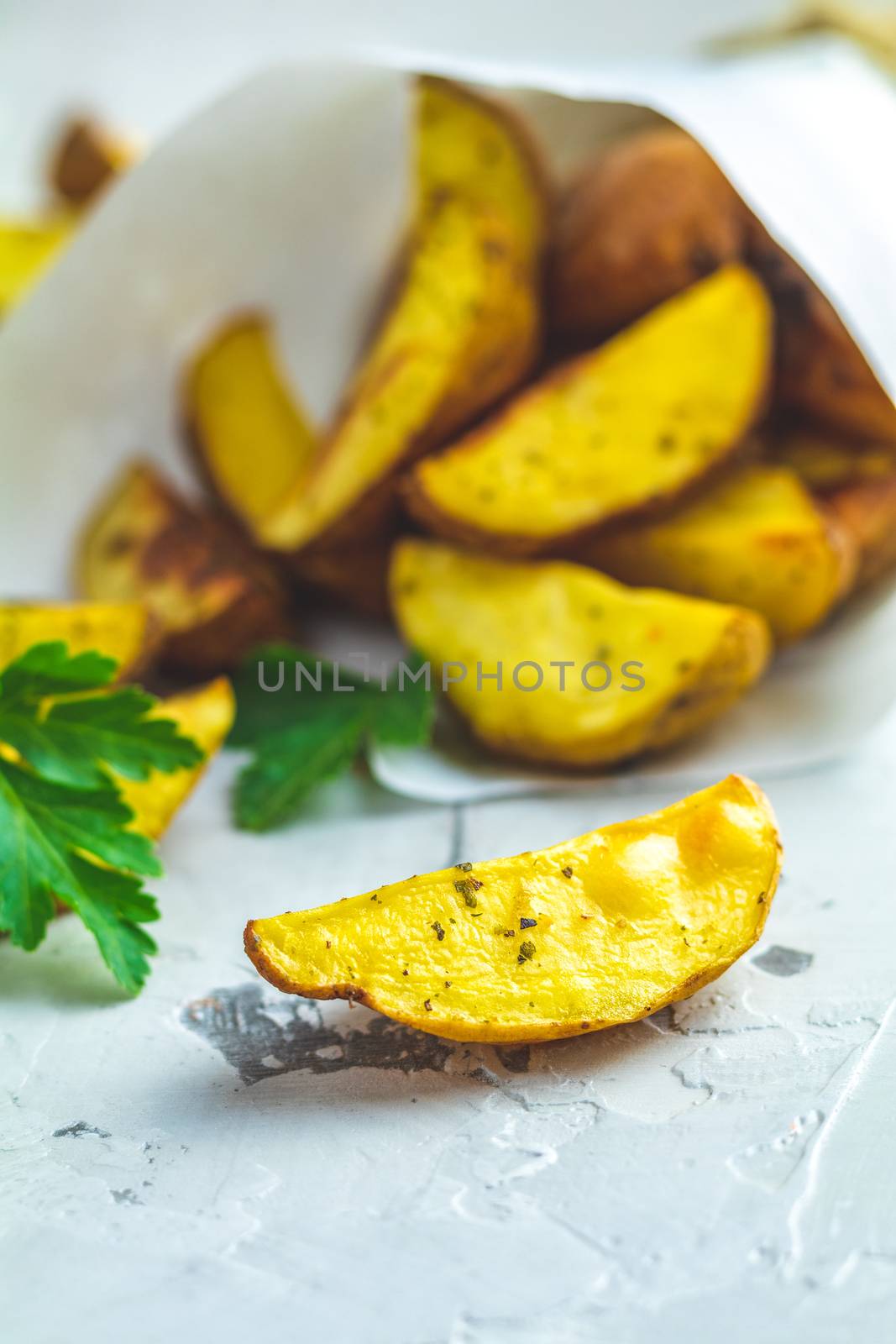 Baked potato wedges on paper with addition sea salt and parsley on a light gray concrete background