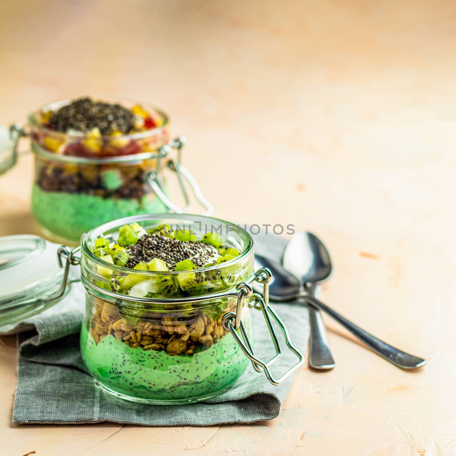 Two chia seed pudding with matcha green tea, kiwi and granola, orange in glasses on light pink concrete background. Healthy breakfast. Square image