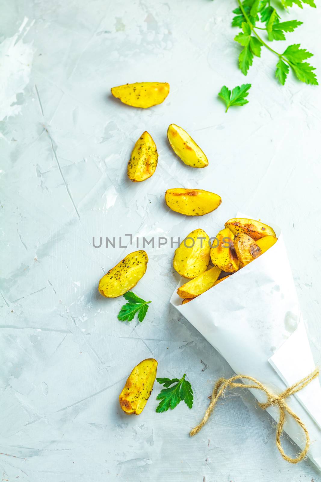 Baked potato wedges on paper with addition sea salt and parsley on a light gray concrete background