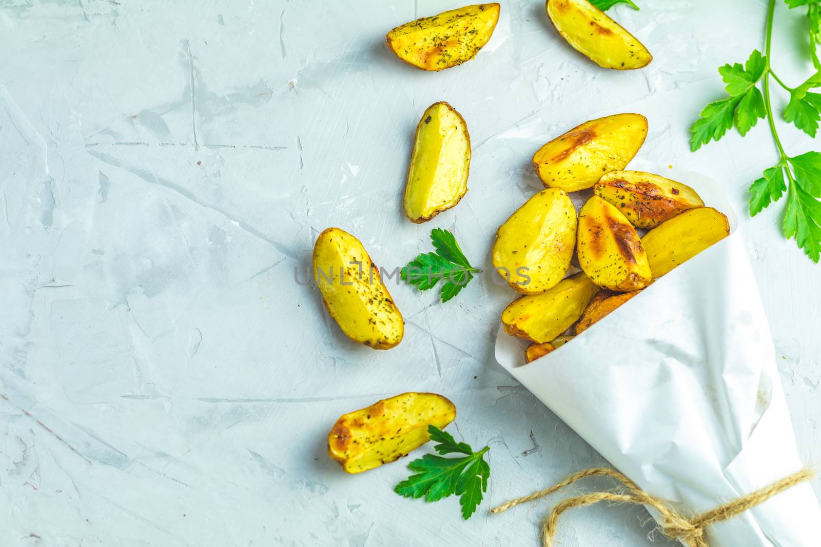 Baked potato wedges on paper with addition sea salt and parsley on a light gray concrete background