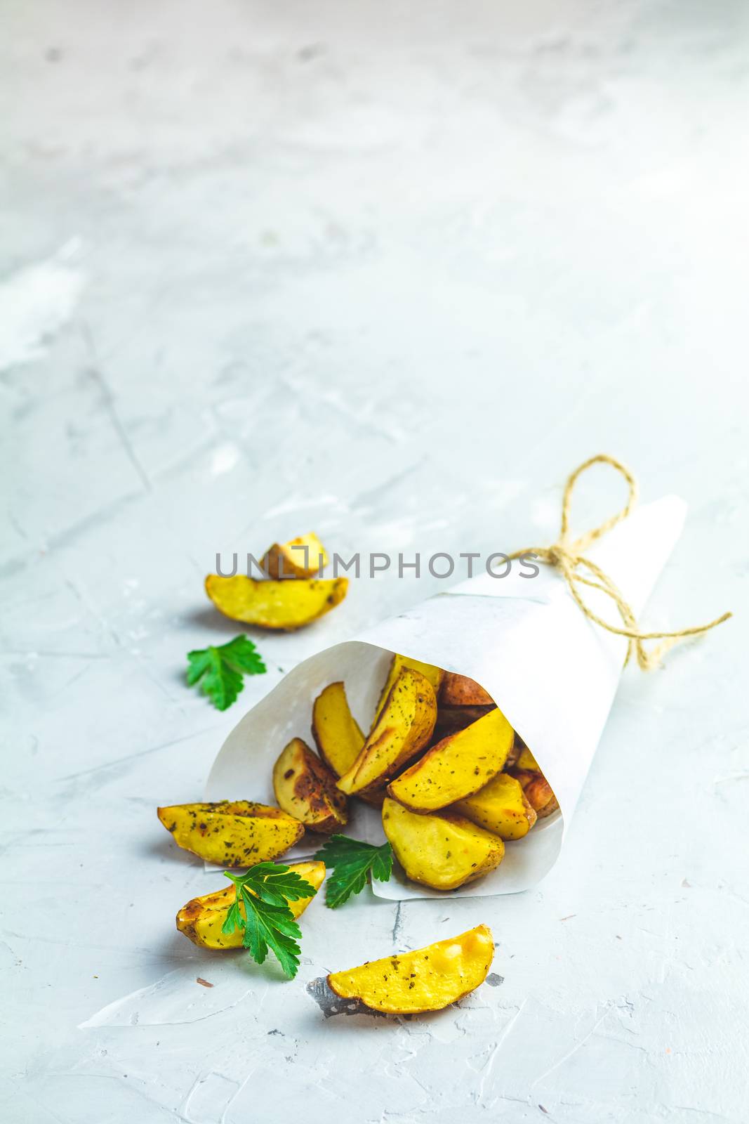 Baked potato wedges on paper with addition sea salt and parsley on a light gray concrete background
