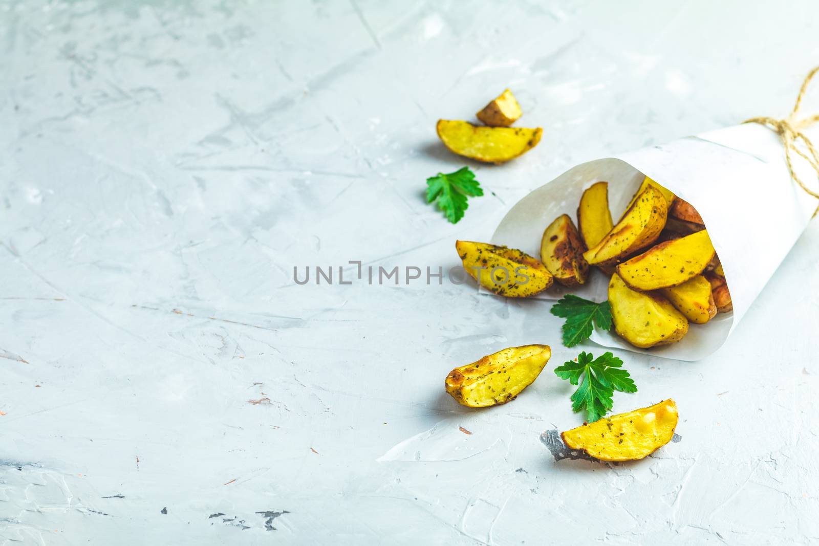 Baked potato wedges on paper with addition sea salt and parsley on a light gray concrete background