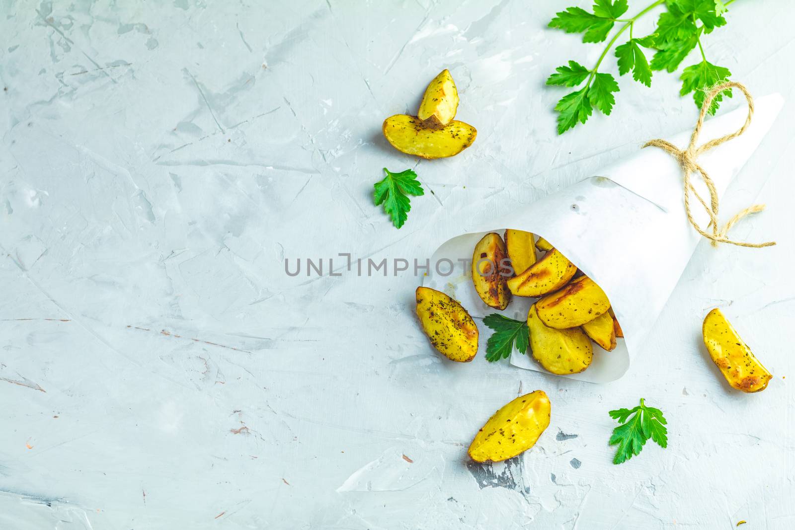 Baked potato wedges on paper with addition sea salt and parsley on a light gray concrete background