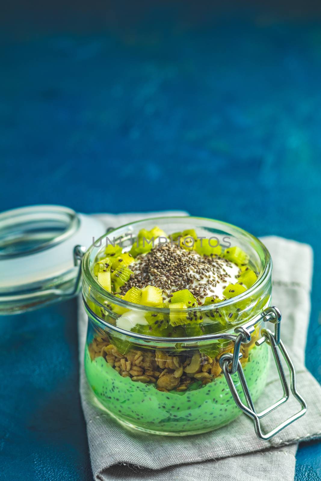 Chia seed pudding with matcha green tea, kiwi and granola in glass on dark blue concrete background. Healthy breakfast.