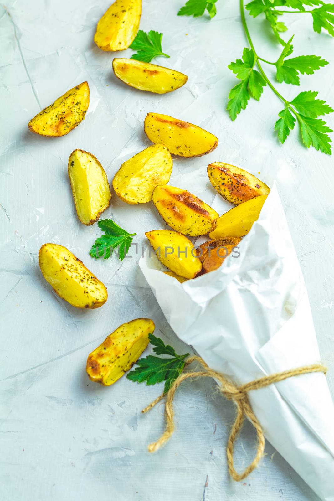 Baked potato wedges on paper with addition sea salt and parsley on a light gray concrete background