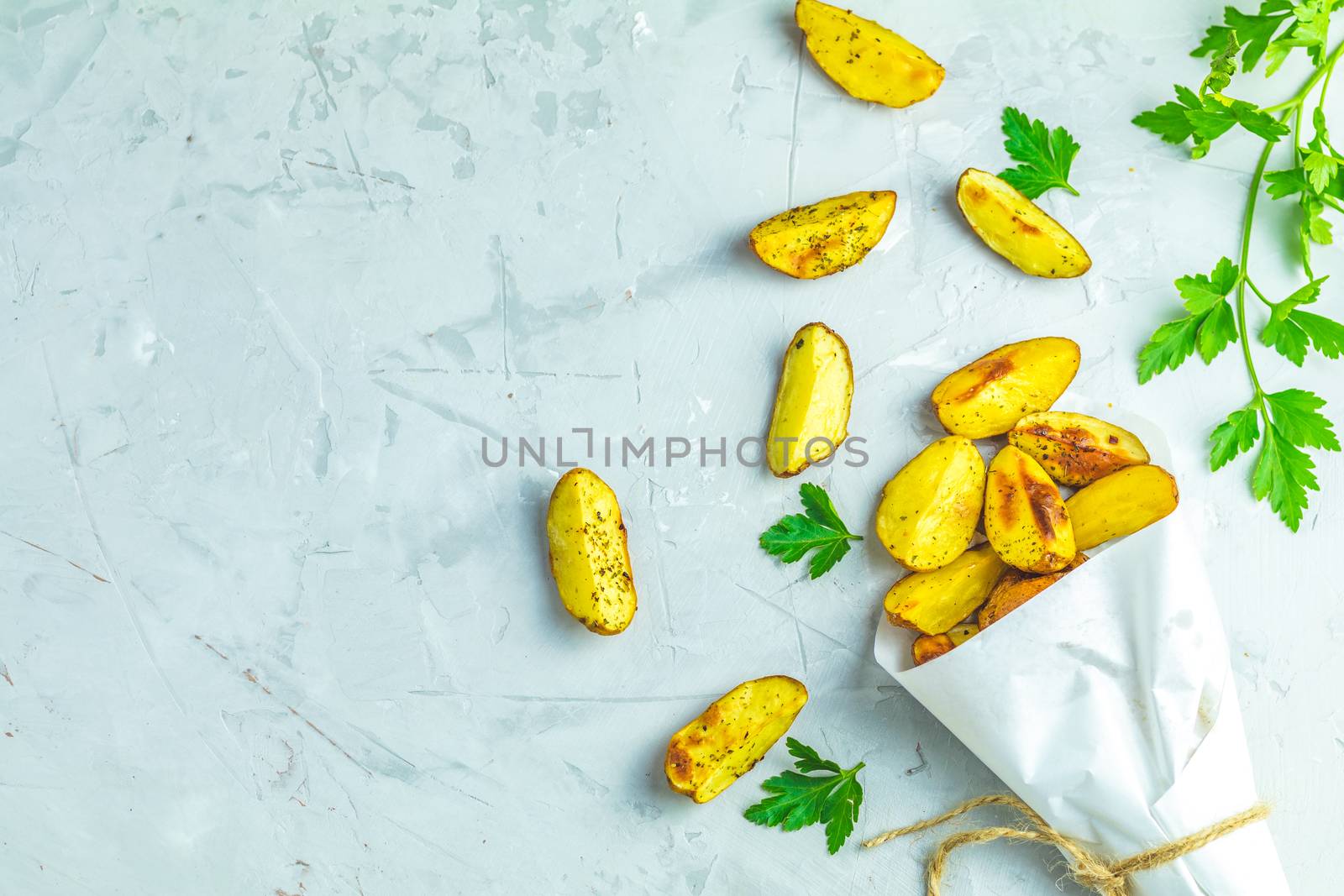 Baked potato wedges on paper with addition sea salt and parsley on a light gray concrete background