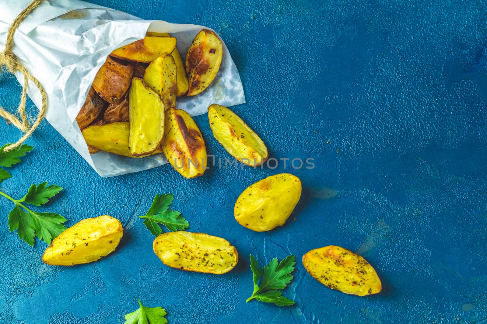 Baked potato wedges on paper with addition sea salt and parsley on a dark blue concrete background