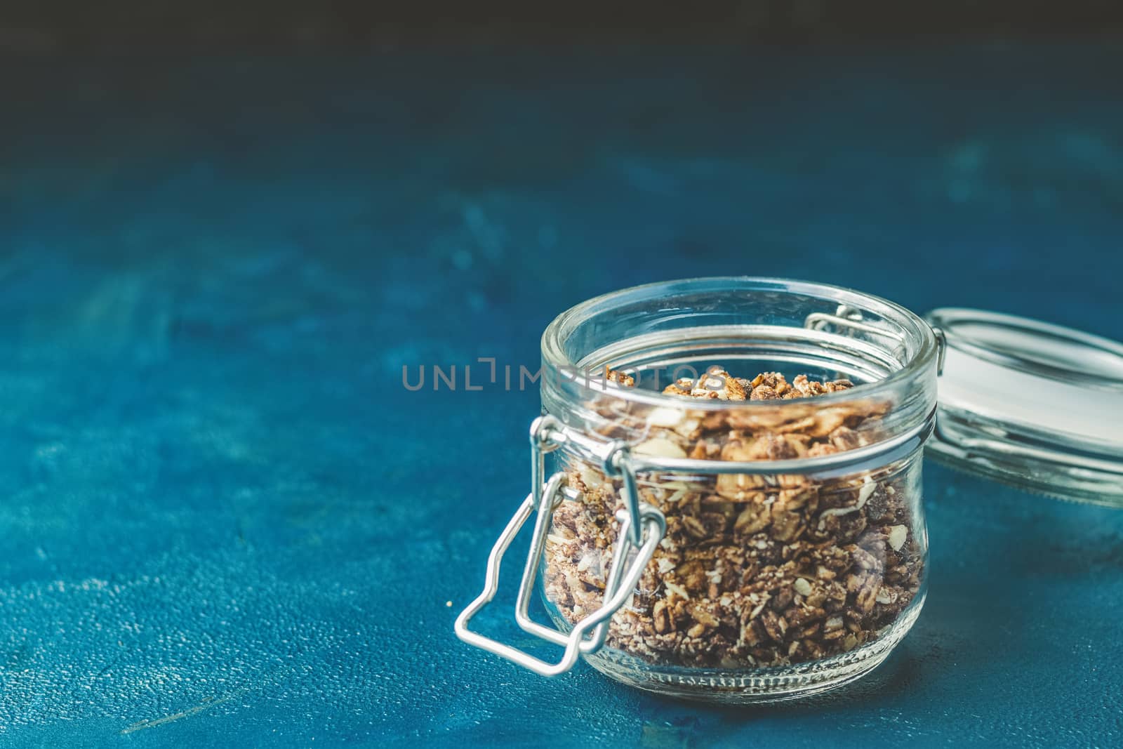 Open glass jar of organic granola with berries, coconut chips and seeds on a dark blue concrete table surface.