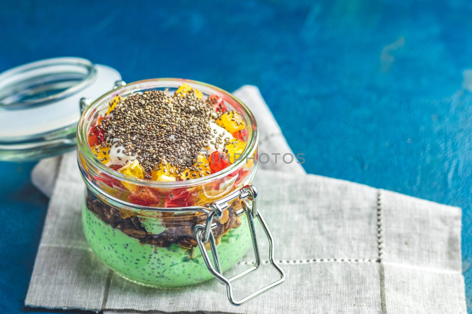 Chia seed pudding with matcha green tea, orange and granola in glass on dark blue concrete background. Healthy breakfast.