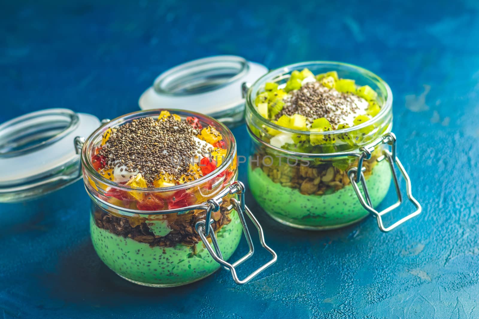 Two chia seed pudding with matcha green tea, kiwi and granola, orange in glasses on dark blue concrete background. Healthy breakfast.