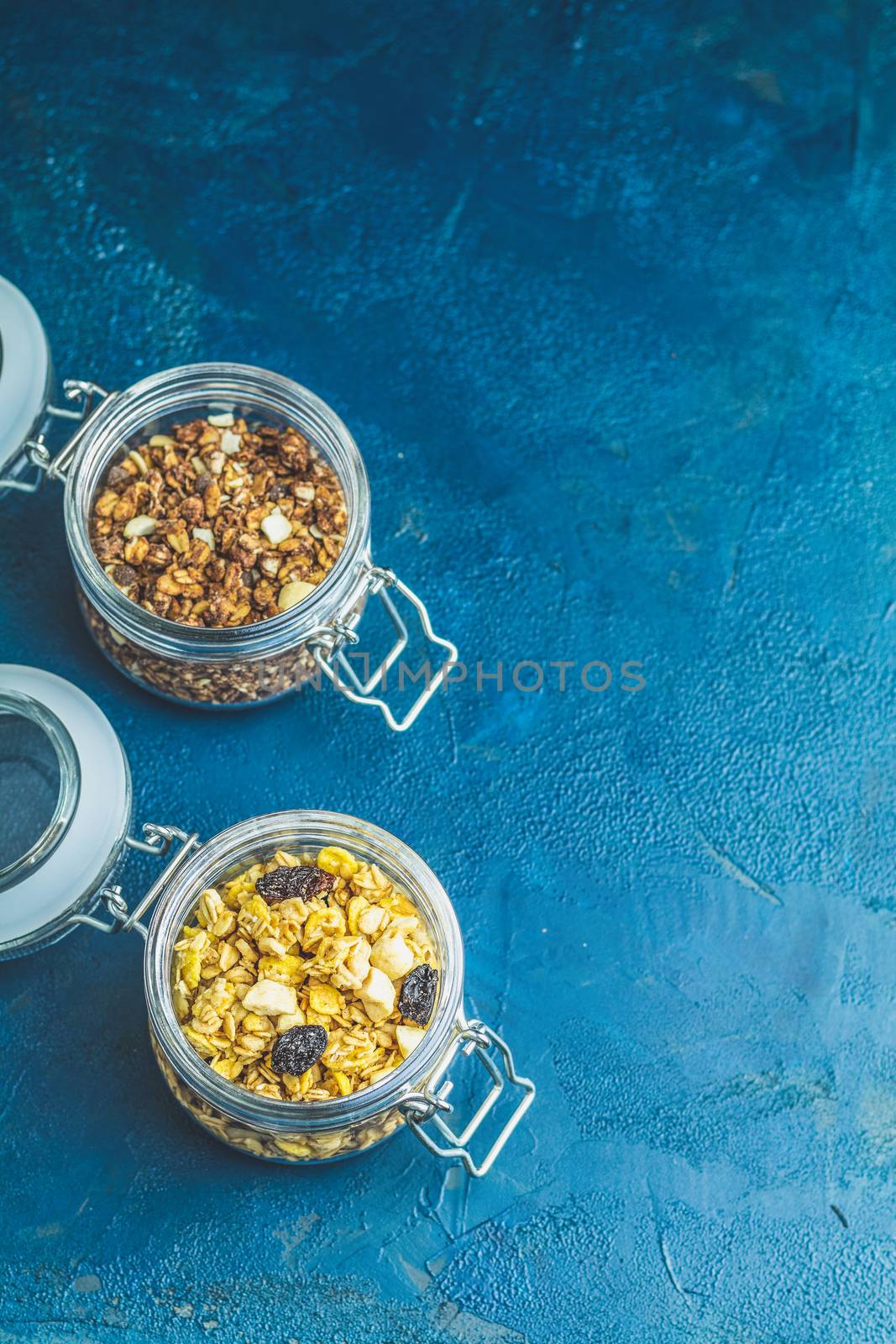 Two open glass jars of organic granola with berries, coconut chips and seeds on a dark blue concrete table surface.