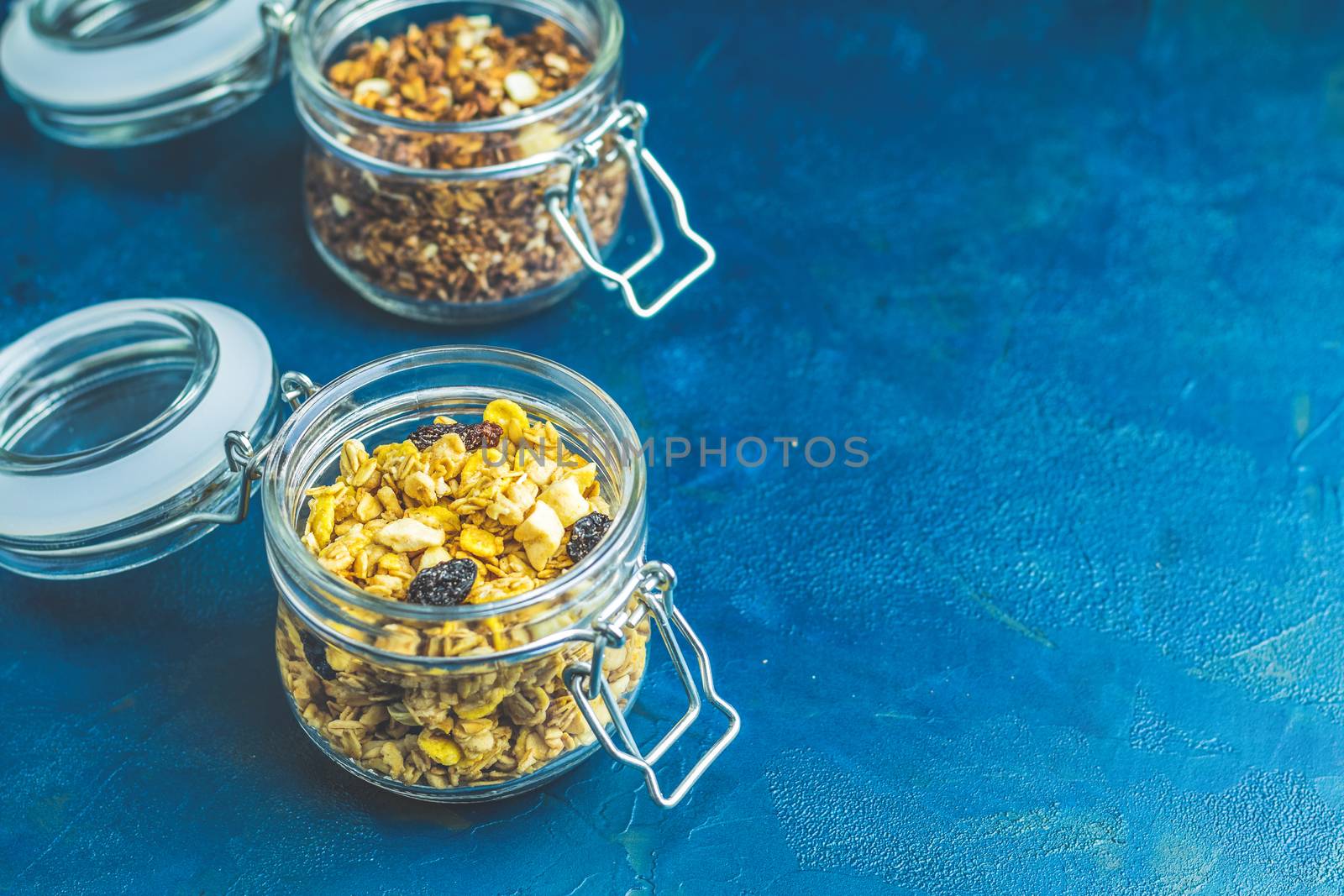 Two open glass jars of organic granola with berries, coconut chips and seeds on a dark blue concrete table surface.