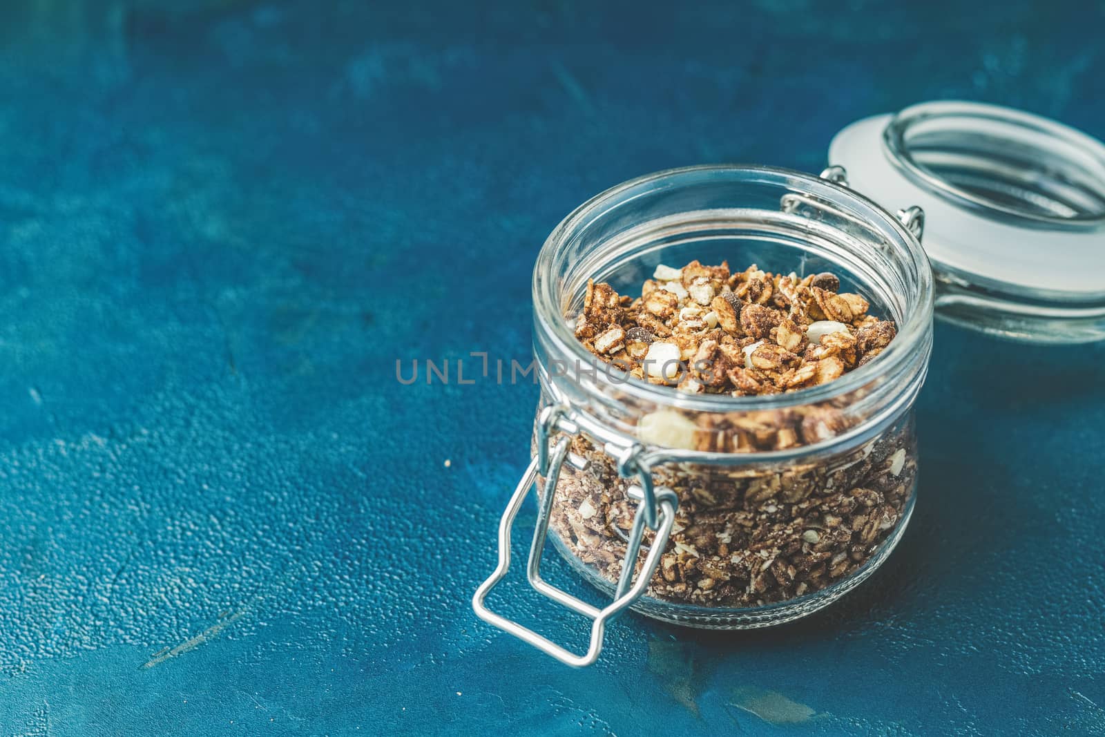 Open glass jar of organic granola with berries, coconut chips and seeds on a dark blue concrete table surface.