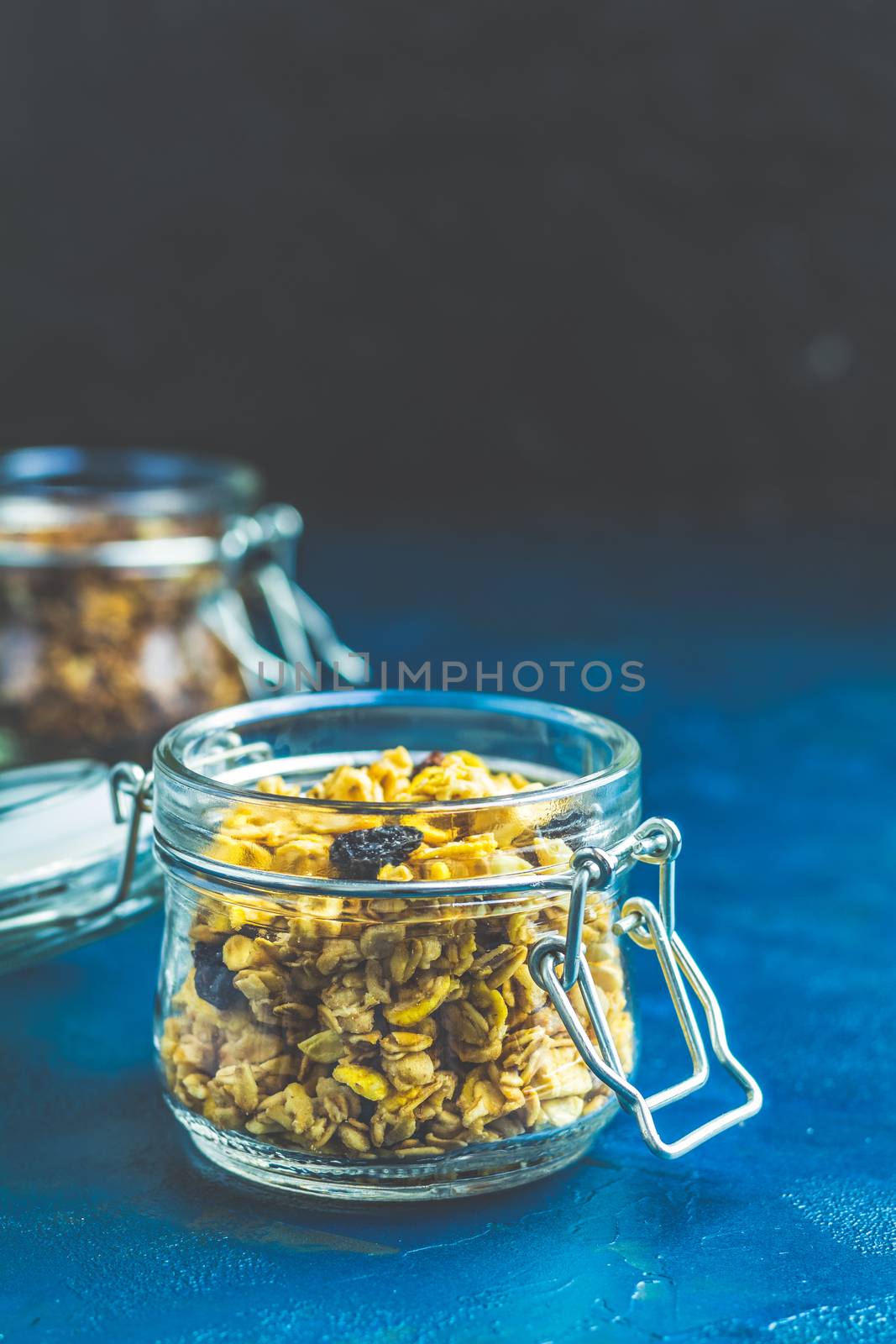 Two open glass jars of organic granola with berries, coconut chips and seeds on a dark blue concrete table surface.