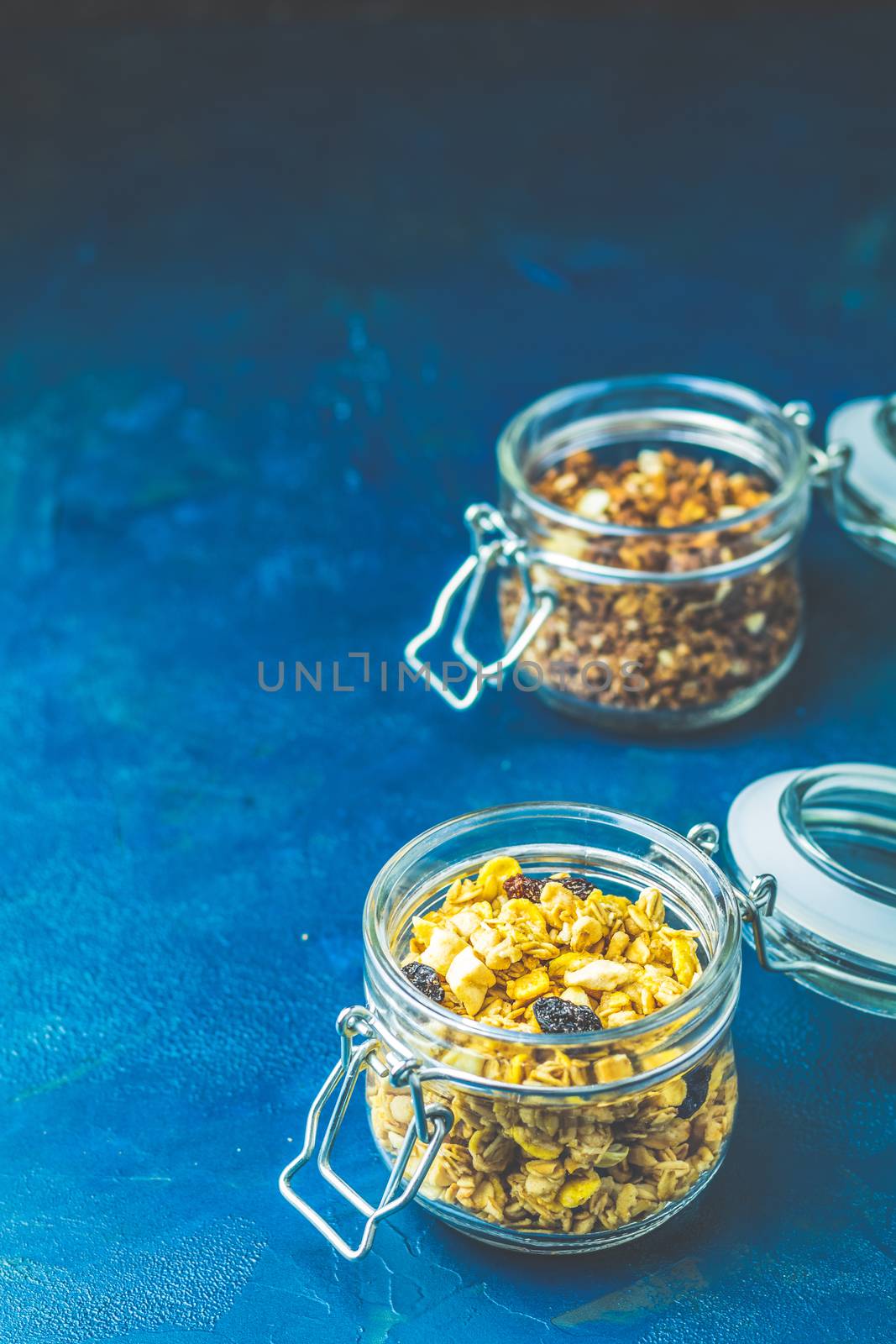 Two open glass jars of organic granola with berries, coconut chips and seeds on a dark blue concrete table surface.