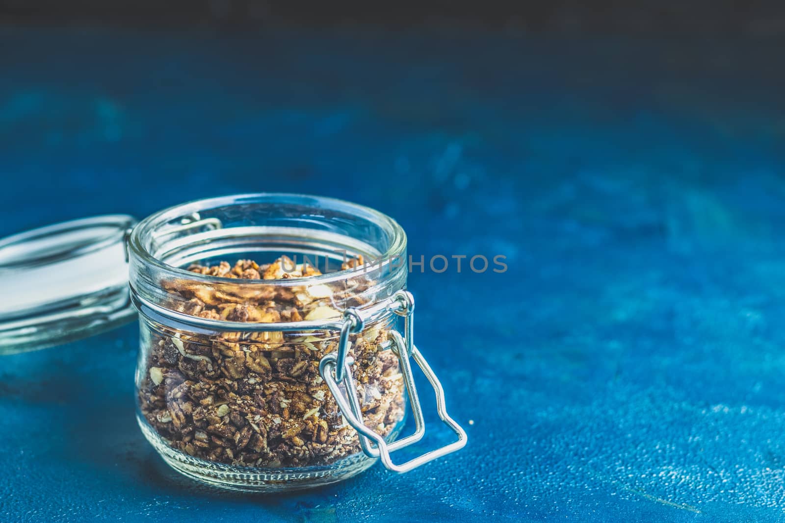 Open glass jar of organic granola with berries, coconut chips and seeds on a dark blue concrete table surface.