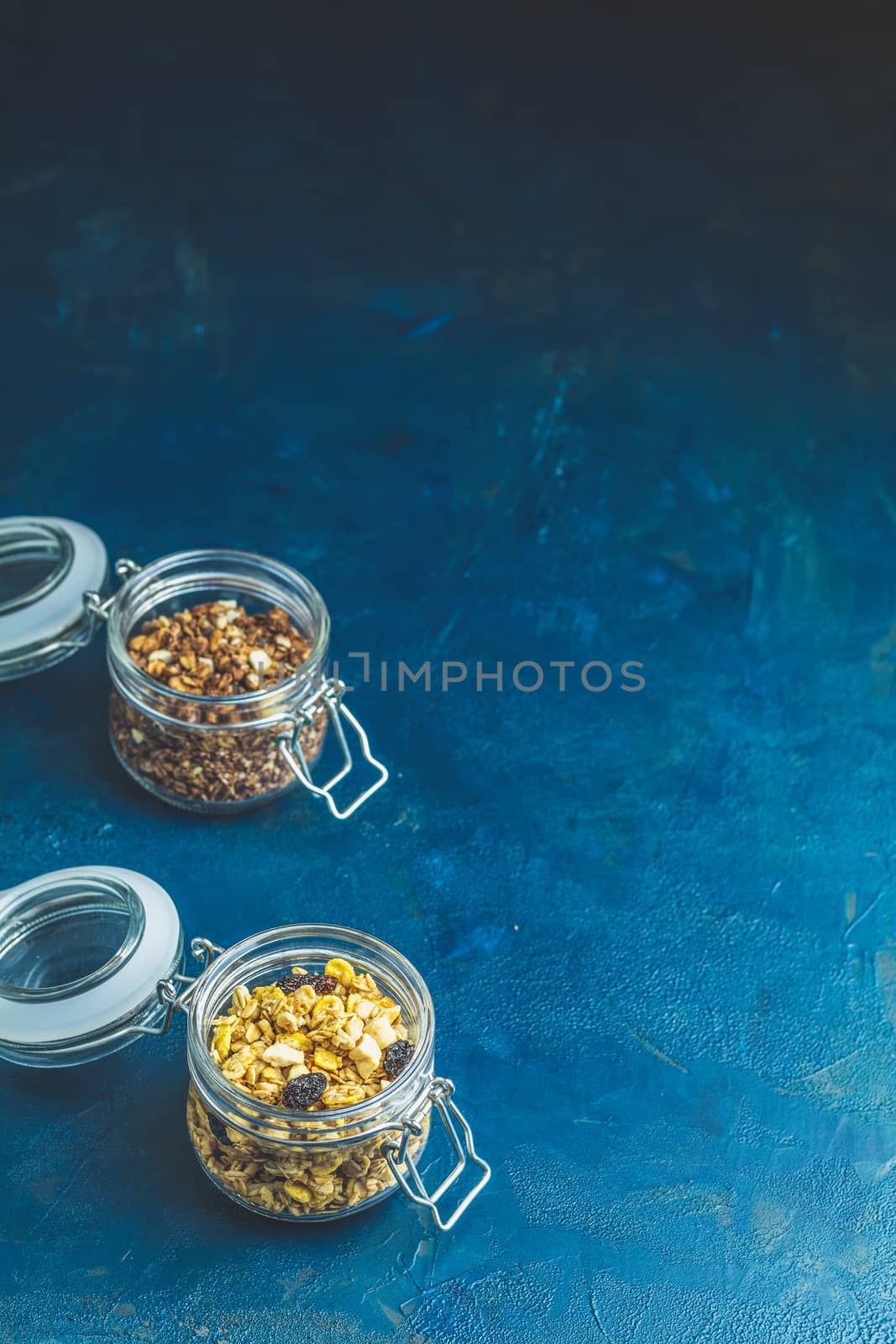 Two open glass jars of organic granola with berries, coconut chips and seeds on a dark blue concrete table surface.