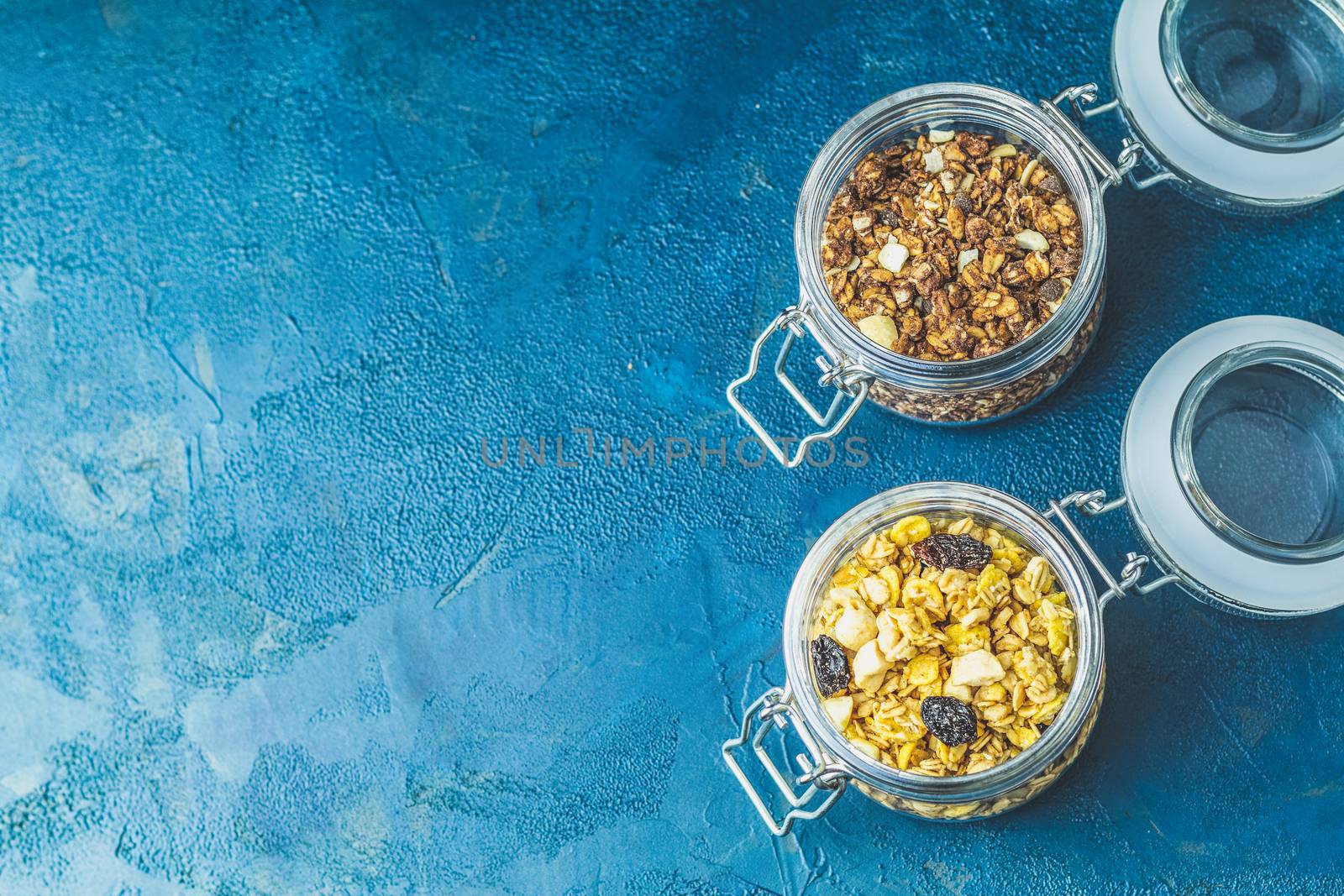 Two open glass jars of organic granola with berries, coconut chips and seeds on a dark blue concrete table surface.