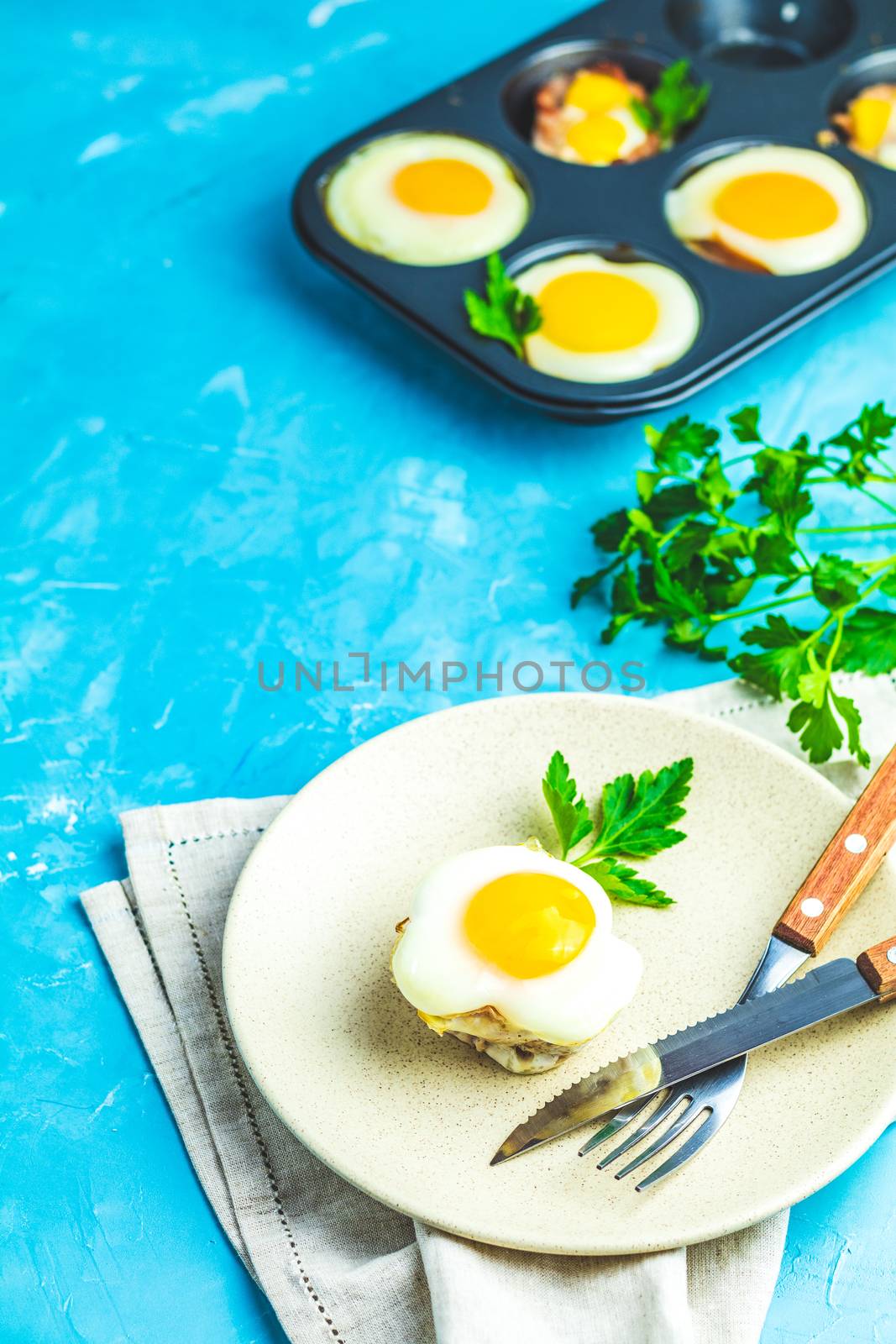 Baked eggs  in light plate and baking molds. Portioned casserole from bacon  and eggs in Italian style. Blue concrete table surface background.