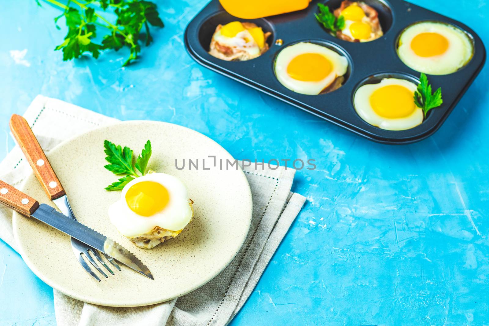 Baked eggs  in light plate and baking molds. Portioned casserole from bacon  and eggs in Italian style. Blue concrete table surface background.