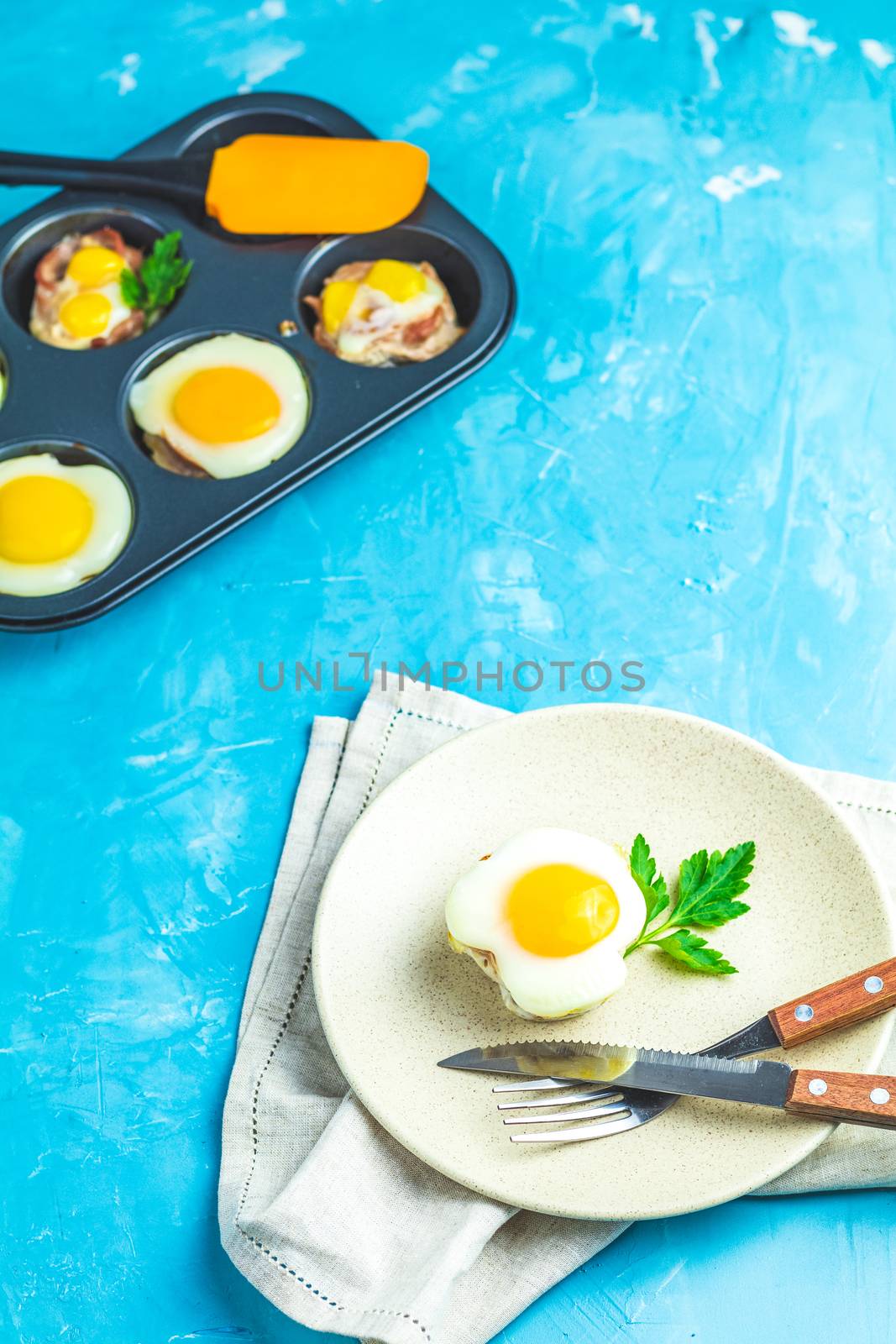 Baked eggs  in light plate and baking molds. Portioned casserole from bacon  and eggs in Italian style. Blue concrete table surface background.