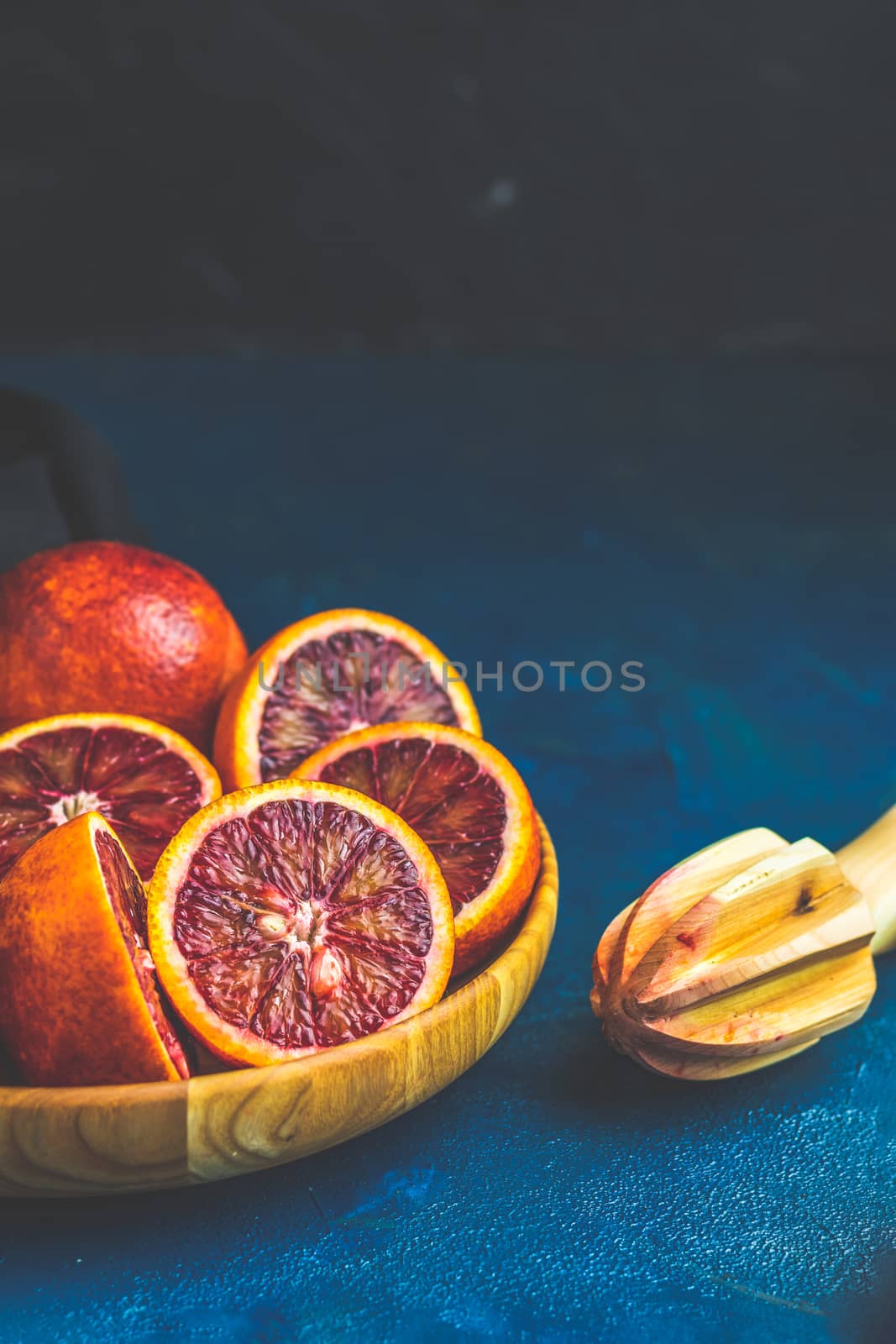 Sliced and whole Sicilian Blood oranges fruits in wooden plate and juicer over dark blue concrete table surface. Dark rustic style.