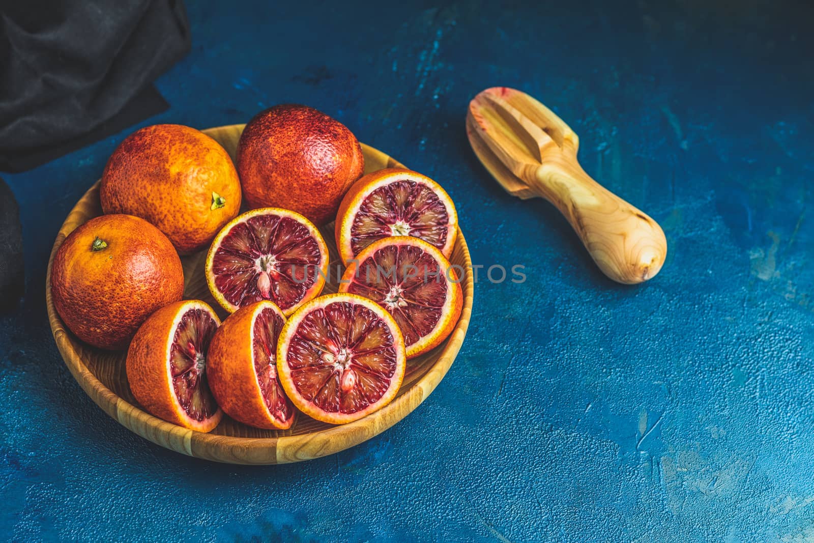 Sliced and whole Sicilian Blood oranges fruits in wooden plate and juicer over dark blue concrete table surface. Dark rustic style.