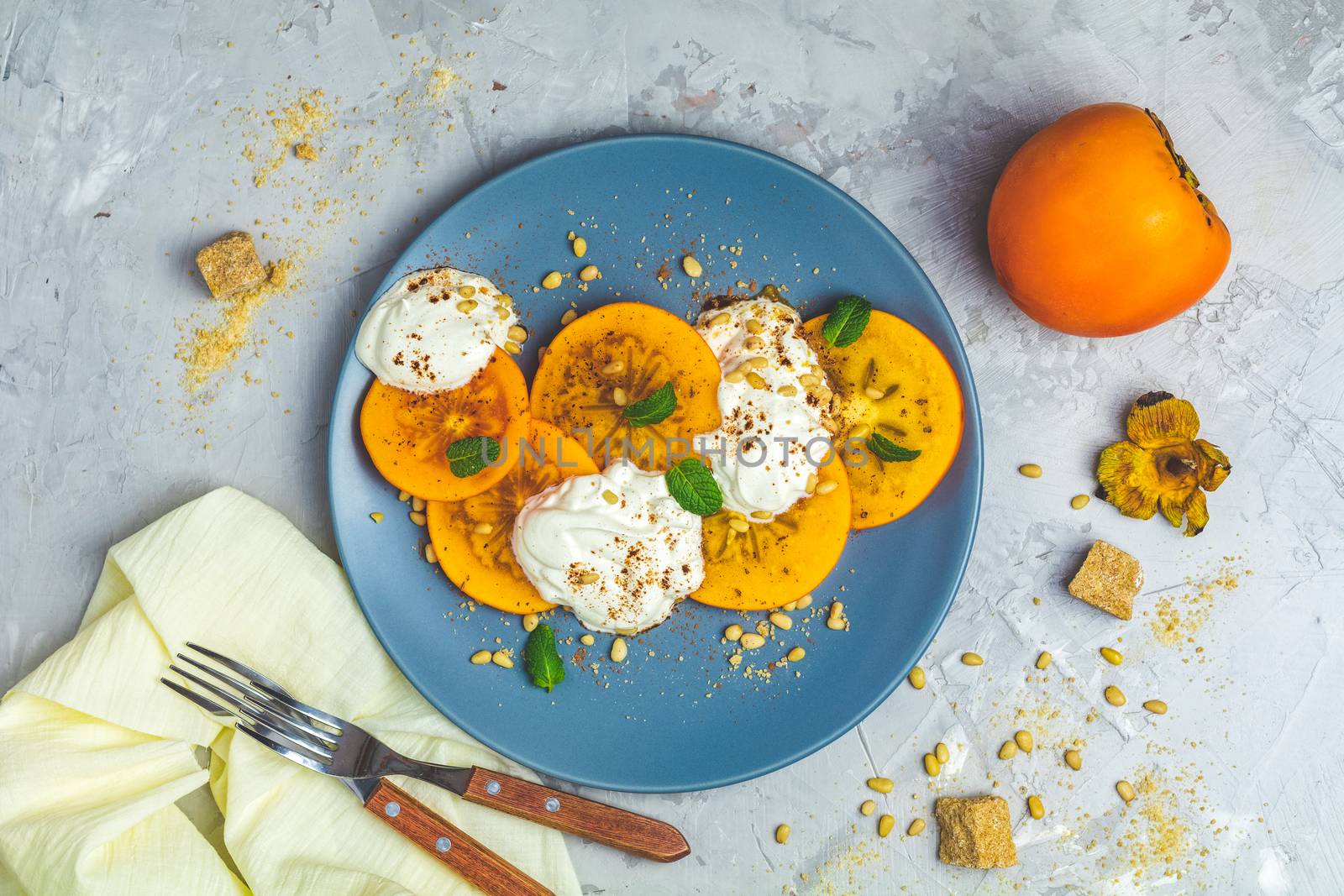 Delicious healthy fruit breakfast. Sliced persimmon with yogurt, brown sugar, pine nuts and fresh mint in blue plate on light gray concrete table surface background, top view, flat lay.