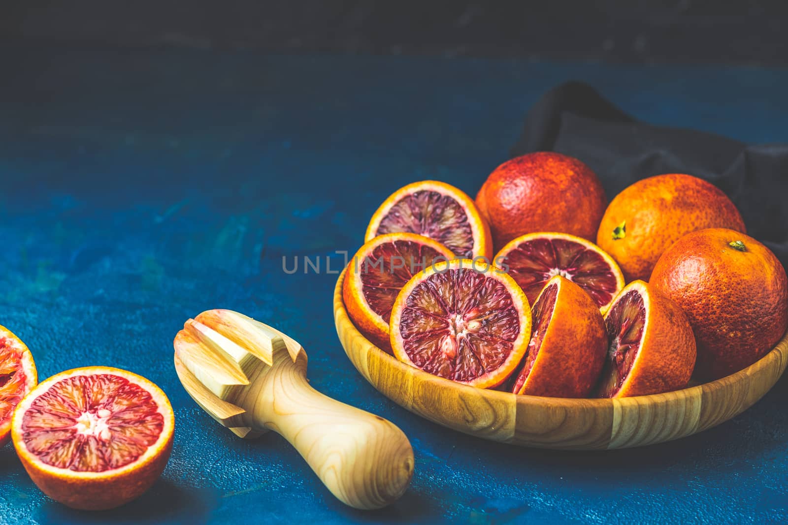 Sliced and whole Sicilian Blood oranges fruits in wooden plate and juicer over dark blue concrete table surface. Dark rustic style.
