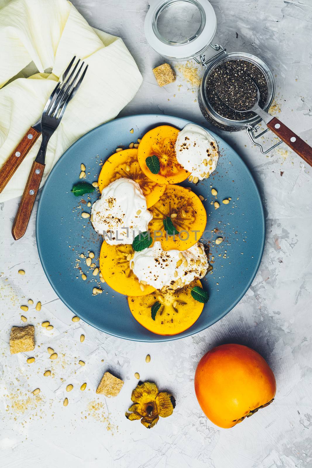 Delicious healthy fruit breakfast. Sliced persimmon with yogurt, chia seeds, brown sugar, pine nuts and fresh mint in blue plate on light gray concrete table surface background, top view, flat lay.