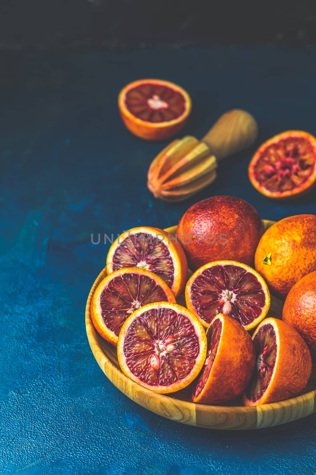 Sliced and whole Sicilian Blood oranges fruits in wooden plate and juicer over dark blue concrete table surface. Dark rustic style.