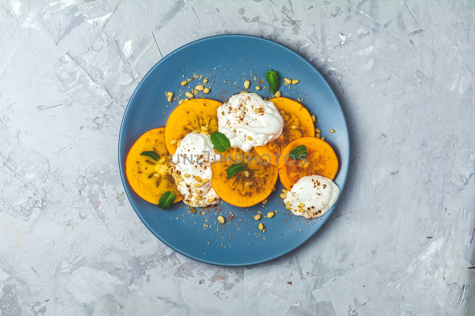 Delicious healthy fruit breakfast. Sliced persimmon with yogurt, brown sugar, pine nuts and fresh mint in blue plate on light gray concrete table surface background, top view, flat lay.