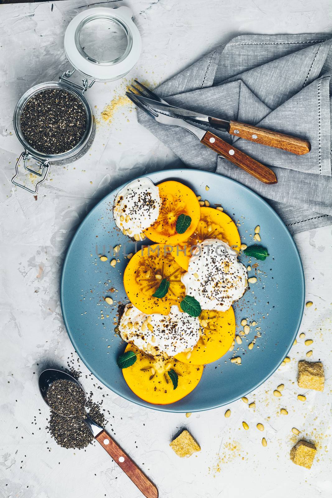 Delicious healthy fruit breakfast. Sliced persimmon with yogurt, chia seeds, brown sugar, pine nuts and fresh mint in blue plate on light gray concrete table surface background, top view, flat lay.