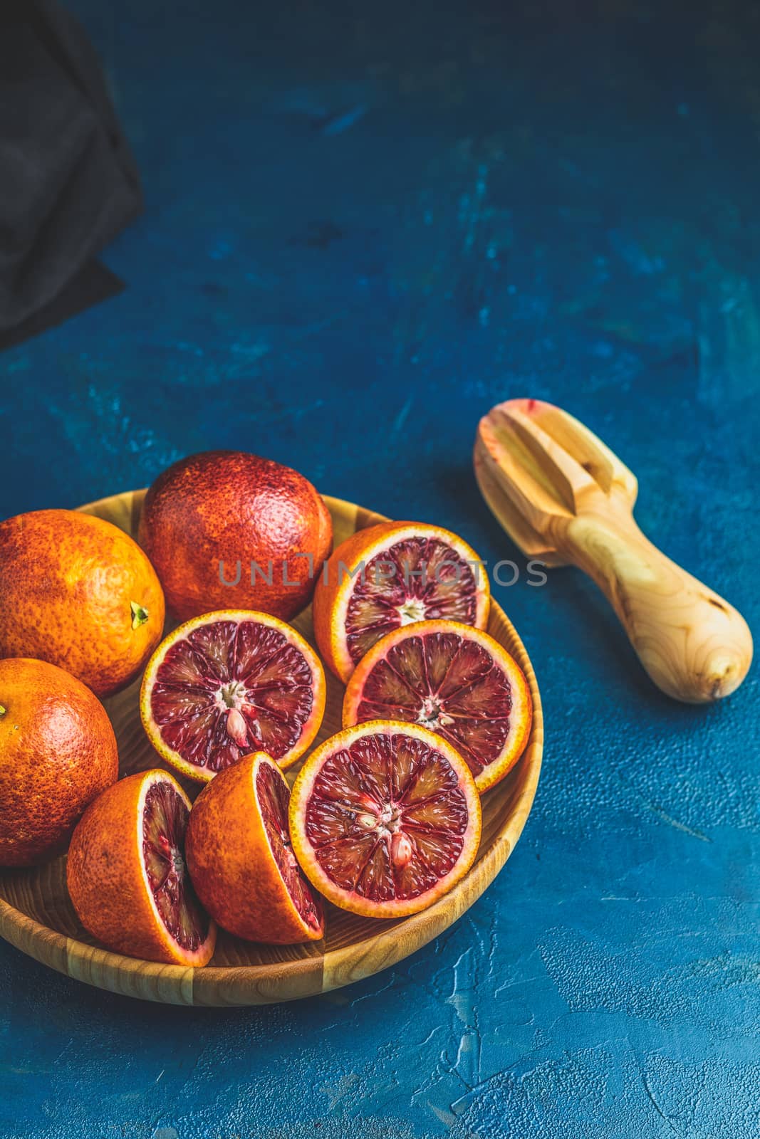 Sliced and whole Sicilian Blood oranges fruits in wooden plate and juicer over dark blue concrete table surface. Dark rustic style.