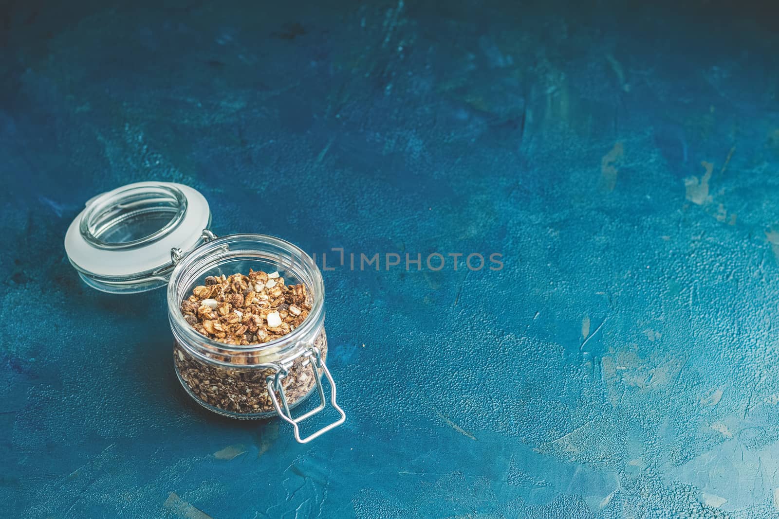 Open glass jar of organic granola with berries, coconut chips and seeds on a dark blue concrete table surface.