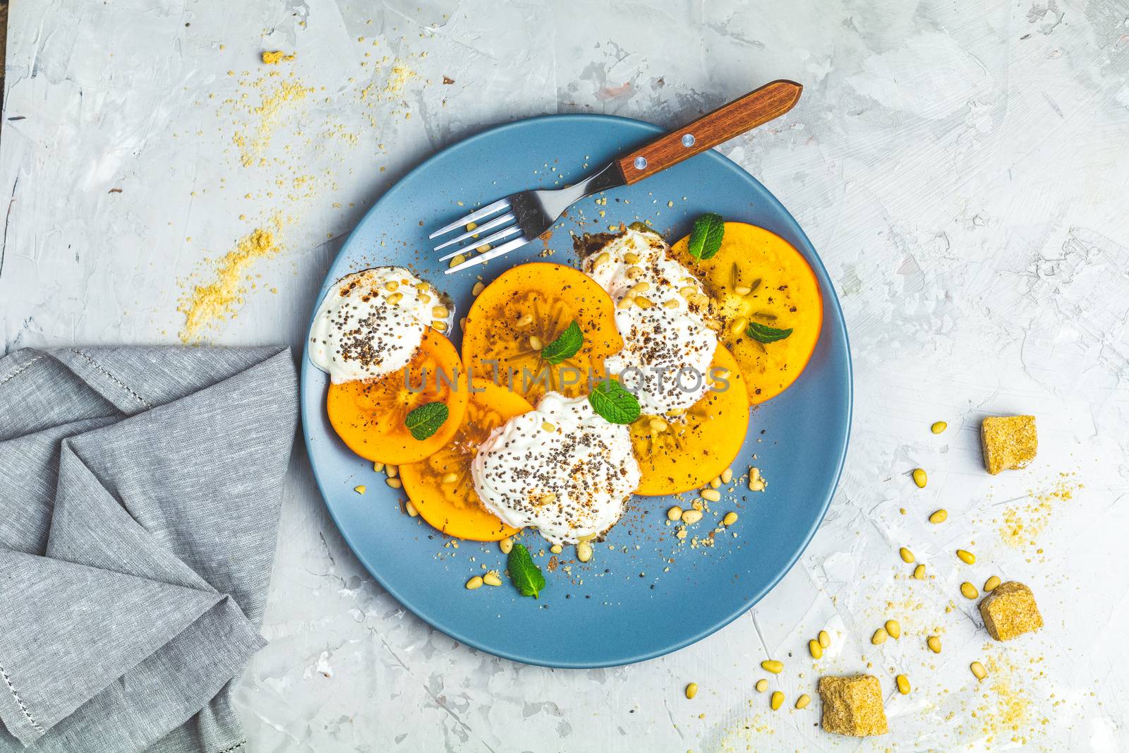 Delicious healthy fruit breakfast. Sliced persimmon with yogurt, chia seeds, brown sugar, pine nuts and fresh mint in blue plate on light gray concrete table surface background, top view, flat lay.