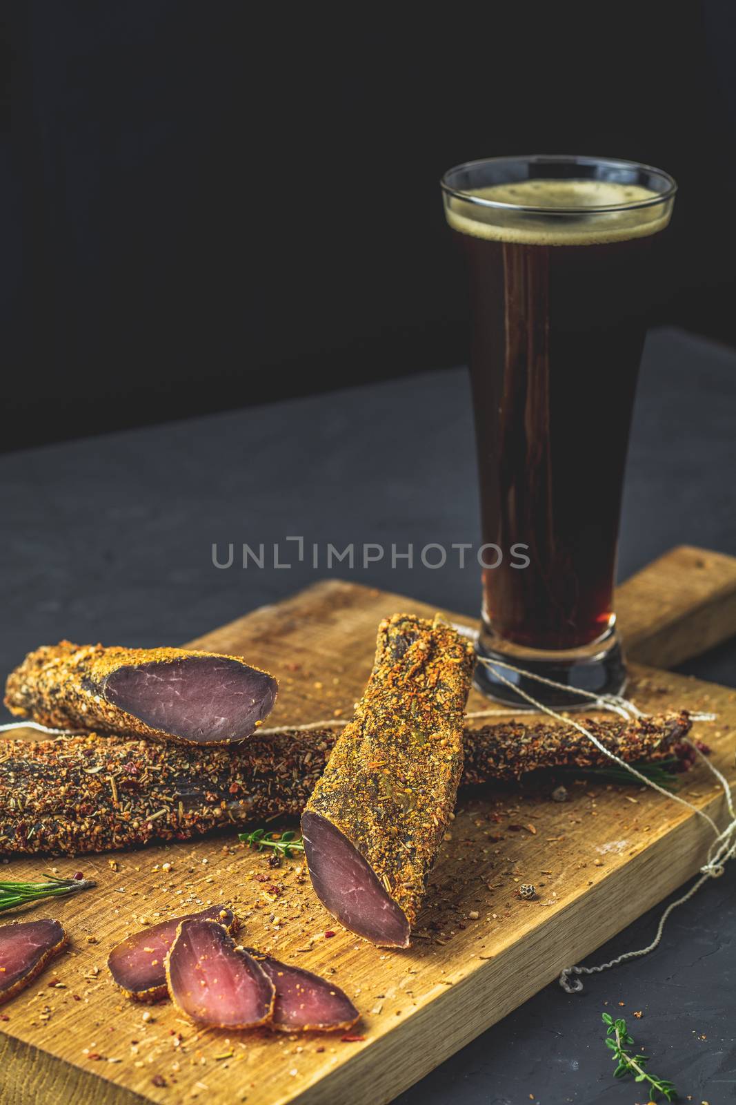 Dark beer in glass and Jerky, basturma, dried meat beef, meat smoked jerky with spices on wooden cutting board, black concrete surface table background.