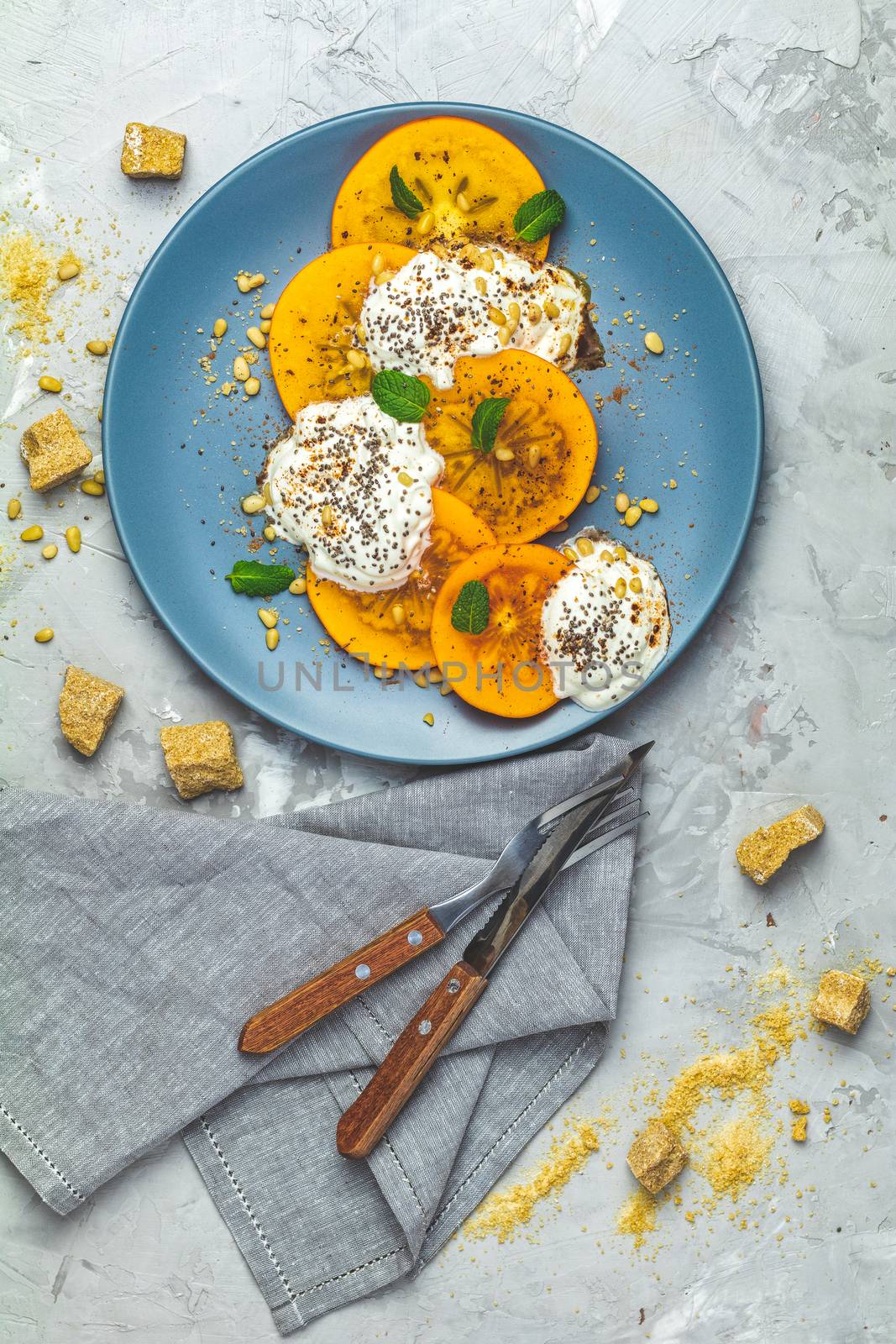 Delicious healthy fruit breakfast. Sliced persimmon with yogurt, chia seeds, brown sugar, pine nuts and fresh mint in blue plate on light gray concrete table surface background, top view, flat lay.