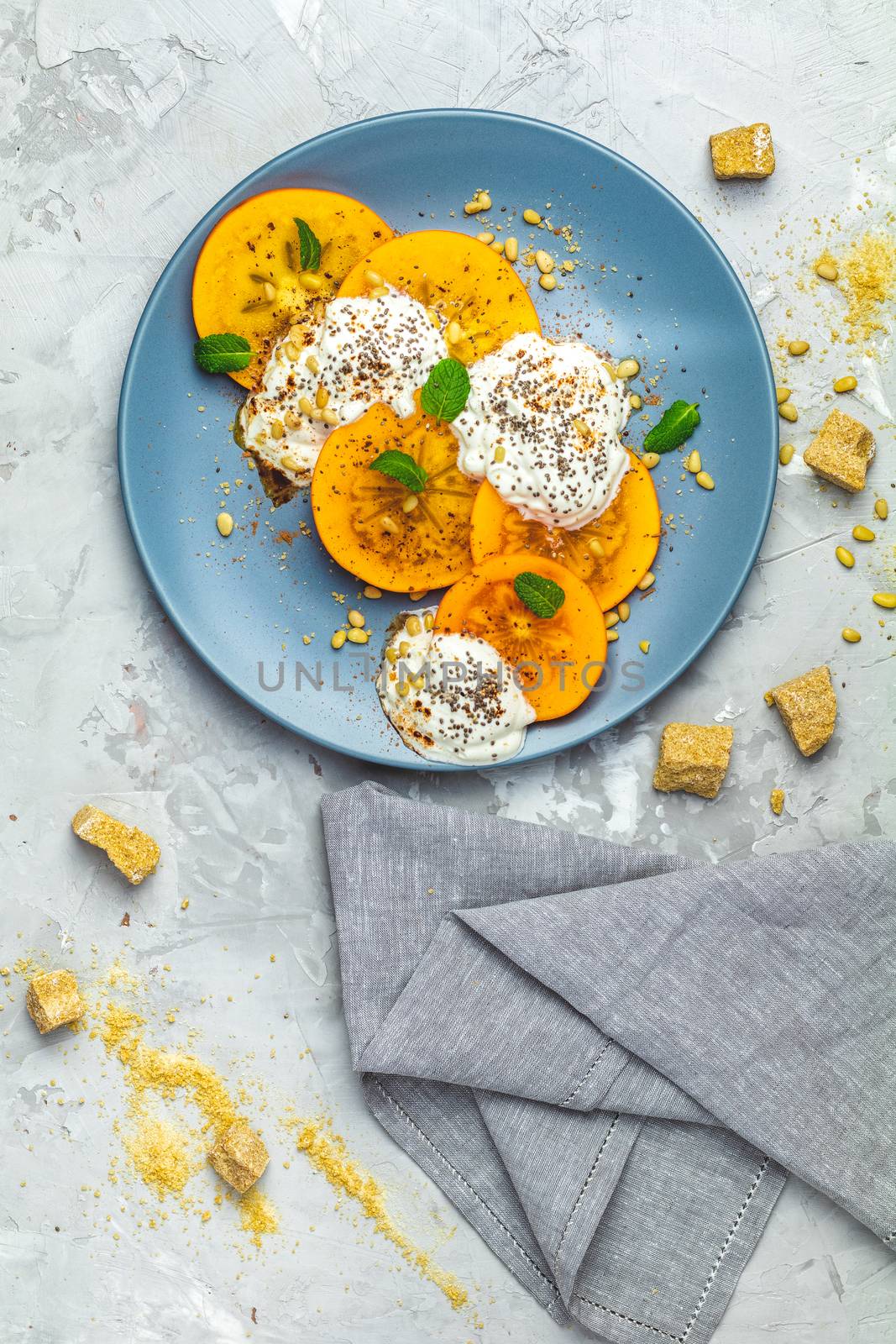 Delicious healthy fruit breakfast. Sliced persimmon with yogurt, brown sugar, pine nuts and fresh mint in blue plate on light gray concrete table surface background, top view, flat lay.