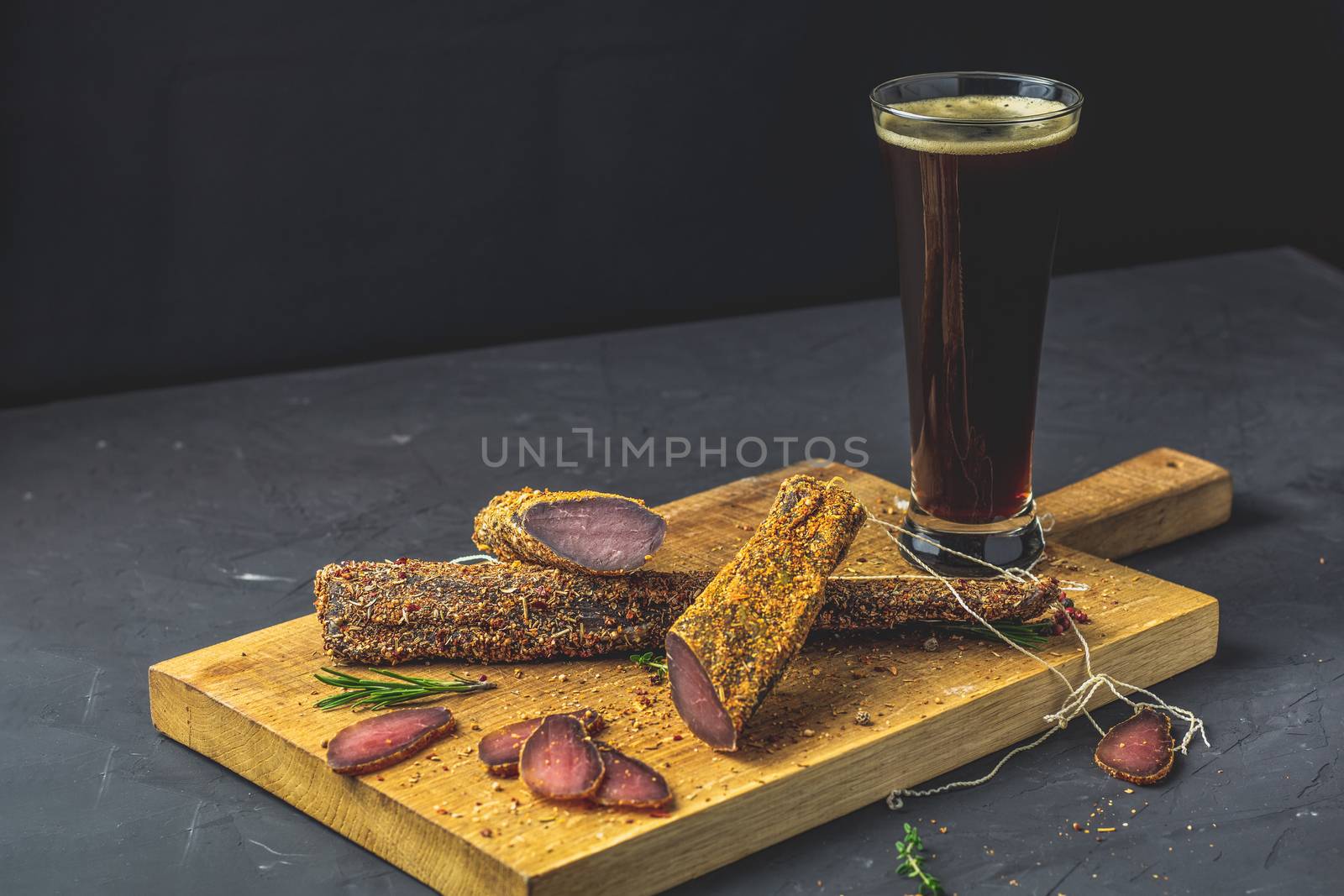 Dark beer in glass and Jerky, basturma, dried meat beef, meat smoked jerky with spices on wooden cutting board, black concrete surface table background.