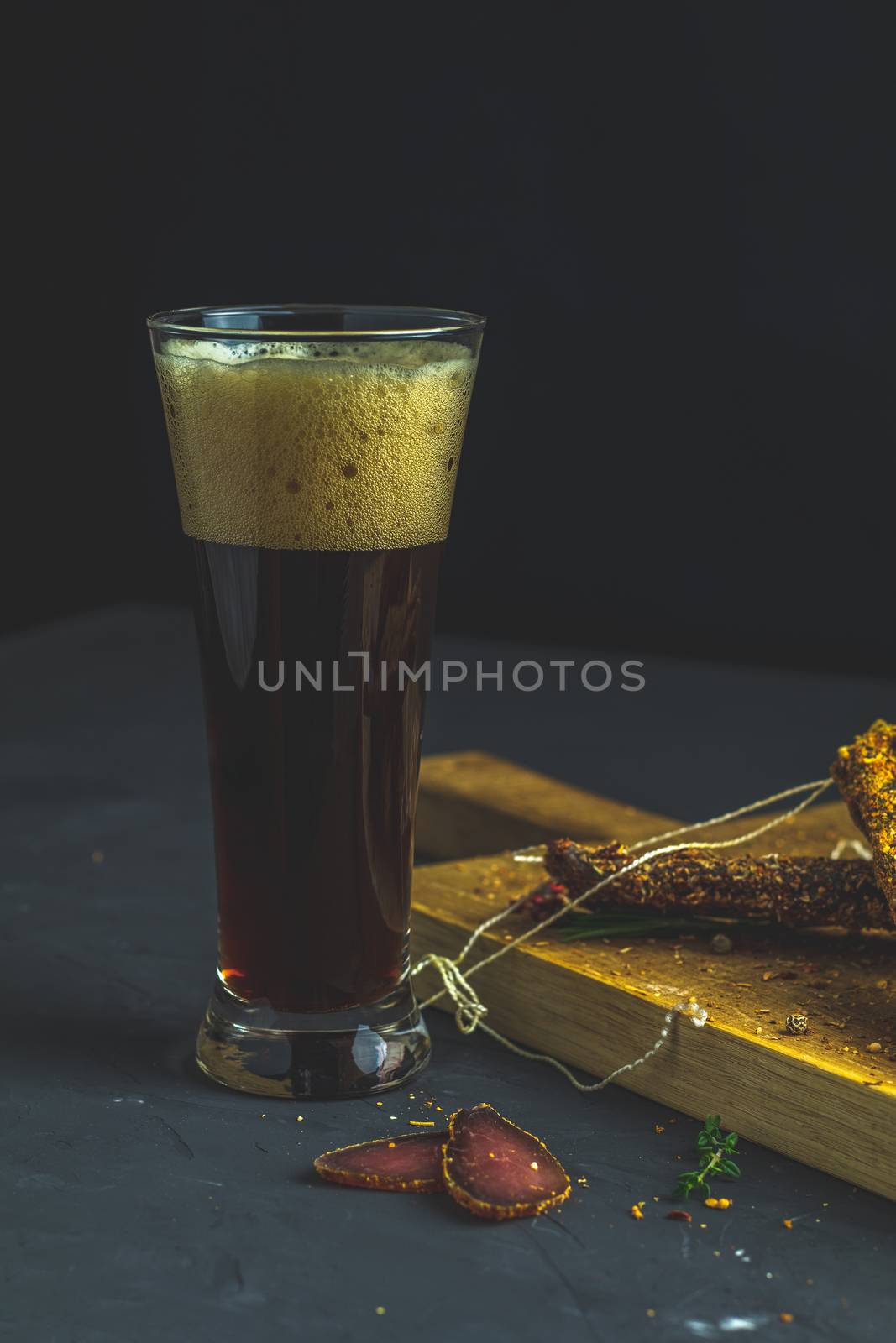 Dark beer in glass and Jerky, basturma, dried meat beef, meat smoked jerky with spices on wooden cutting board, black concrete surface table background.