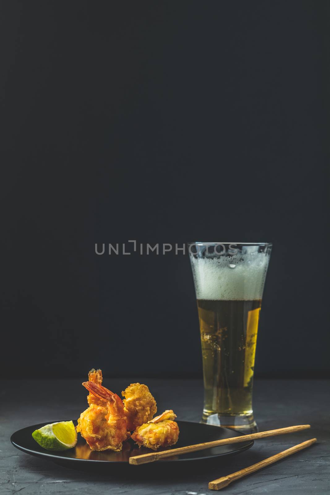 Fried Shrimps tempura with lime in black plate and glass of light beer on dark concrete surface background. Copy space. Seafood tempura dish served japanese or eastern Asia style with chopsticks