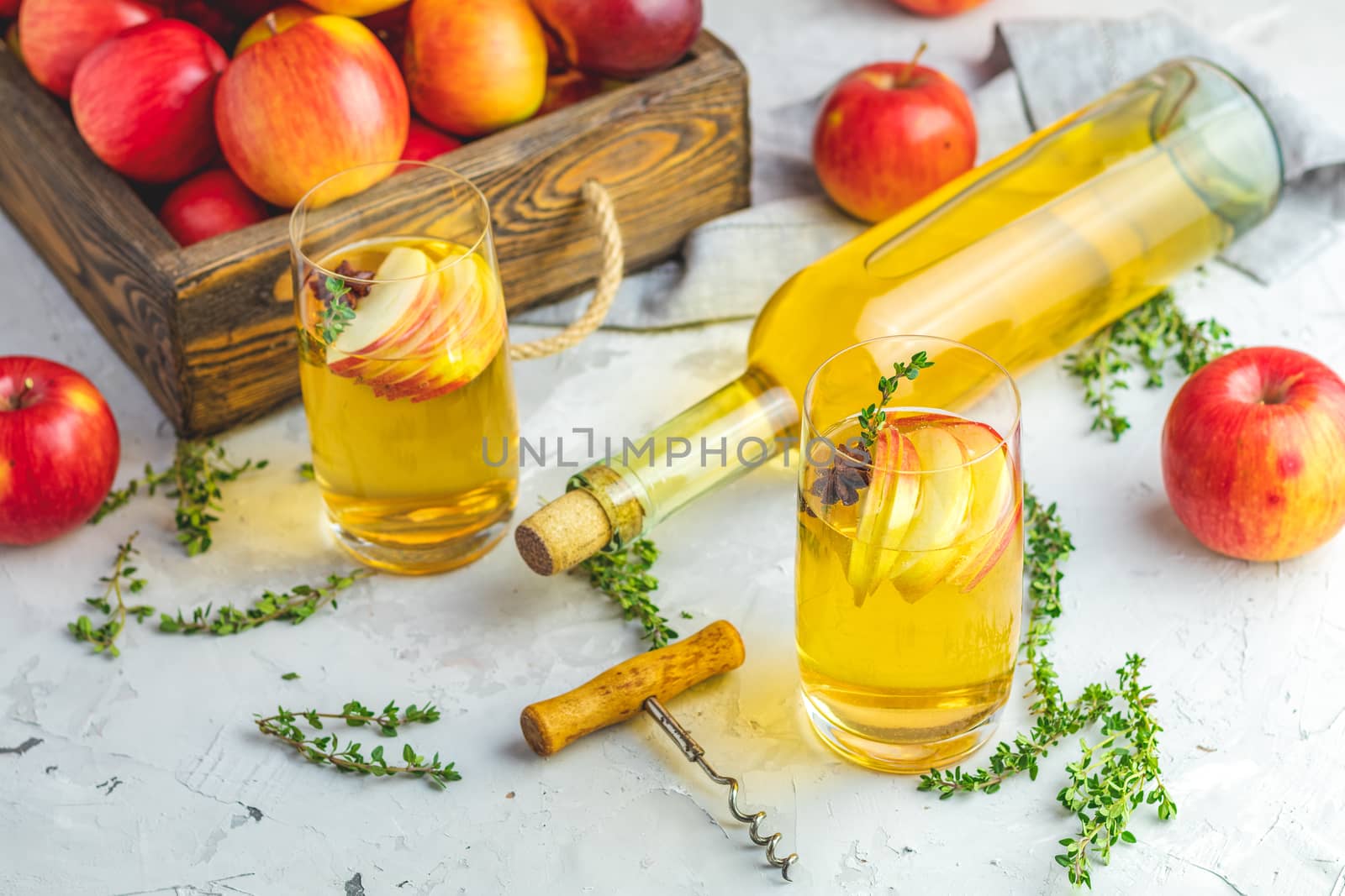 Hard apple cider cocktail with fall fresh thyme and star anise. Bottle and glasses of homemade organic apple cider with fresh apples in box, light concrete table surface. Shallow depth of the field.