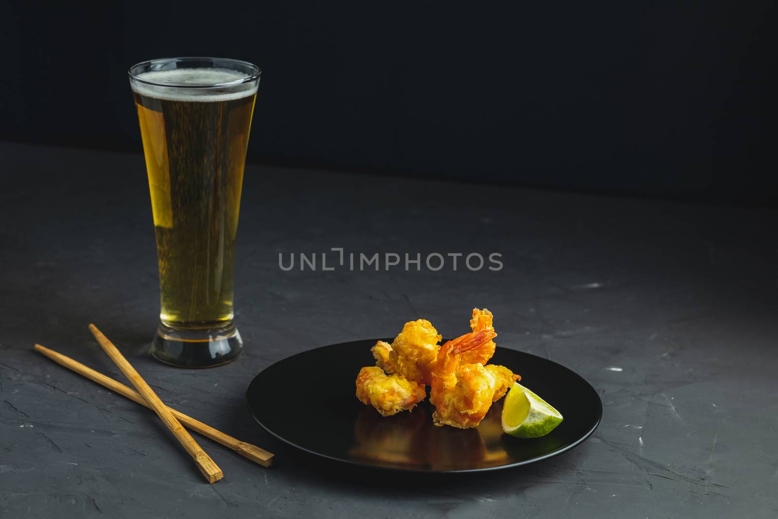 Fried Shrimps tempura with lime in black plate and glass of light beer on dark concrete surface background. Copy space. Seafood tempura dish served japanese or eastern Asia style with chopsticks