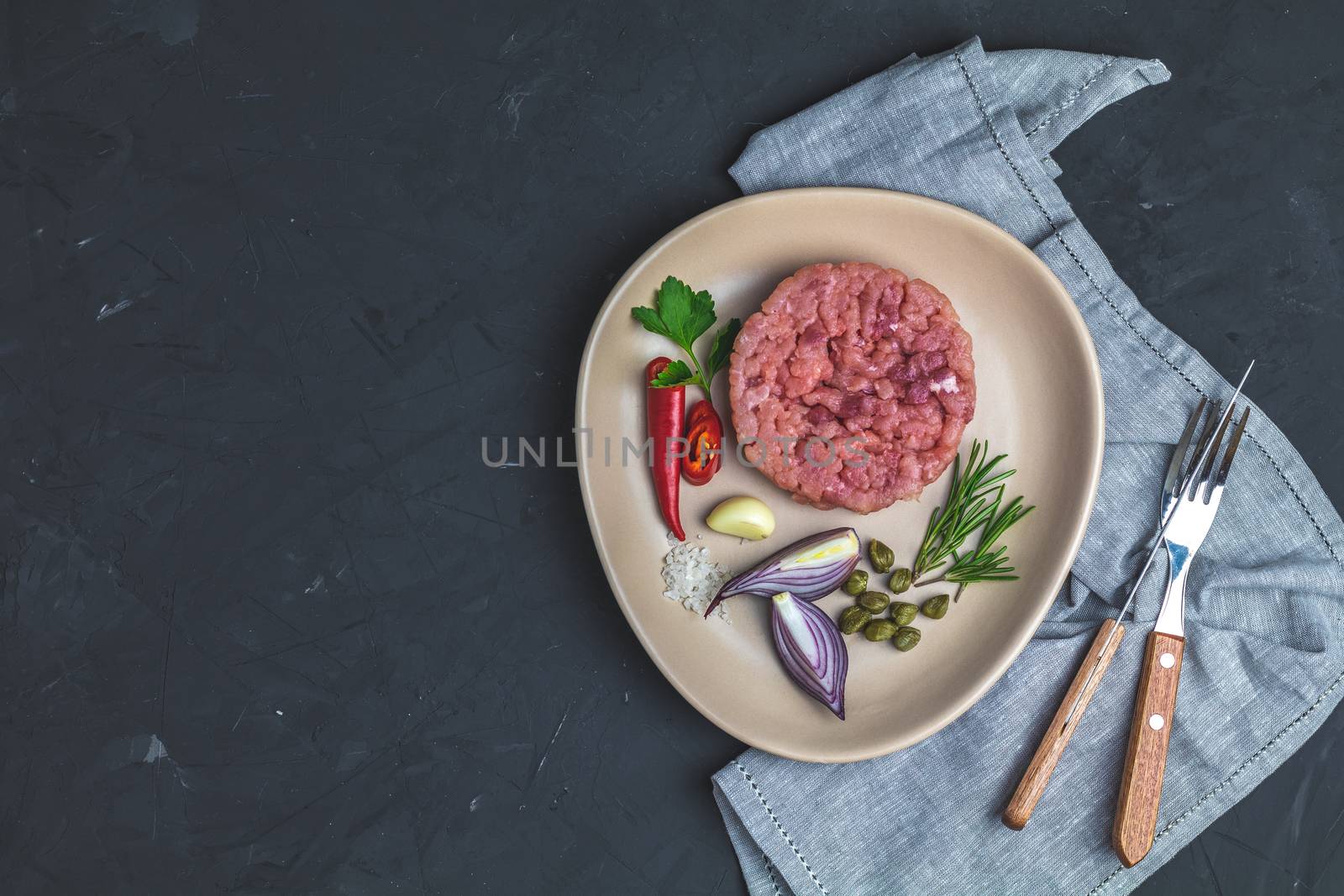 Delicious steak tartare and ingredients on ceramic plate, set of cutlery knife, fork on black stone concrete textured surface background. Copy space background, top view flat lay.