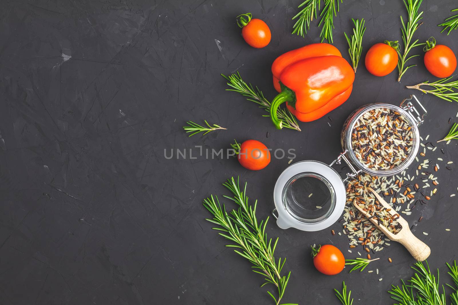 Black, purple, and white rice (Oryza sativa) mix in glass jar on black stone concrete textured surface background. Raw pepper, tomatoes, olive oil and rosemary bunch. Top view with copy space for your text.