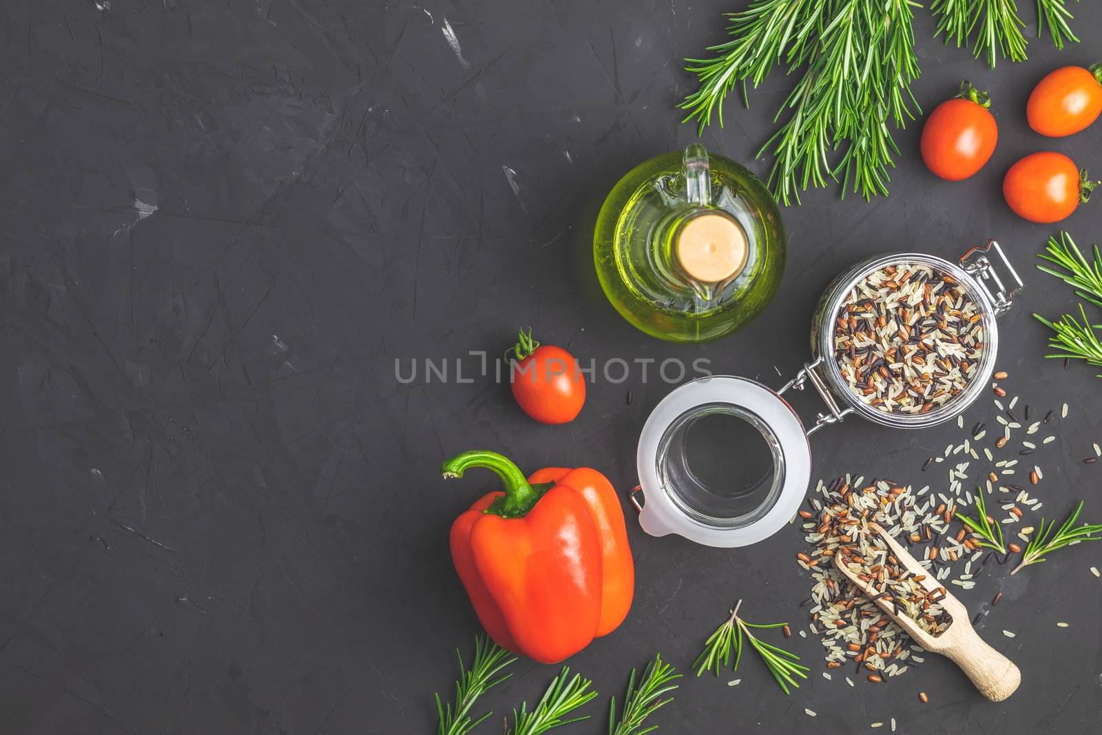 Black, purple, and white rice (Oryza sativa) mix in glass jar on black stone concrete textured surface background. Raw pepper, tomatoes, olive oil and rosemary bunch. Top view with copy space for your text.