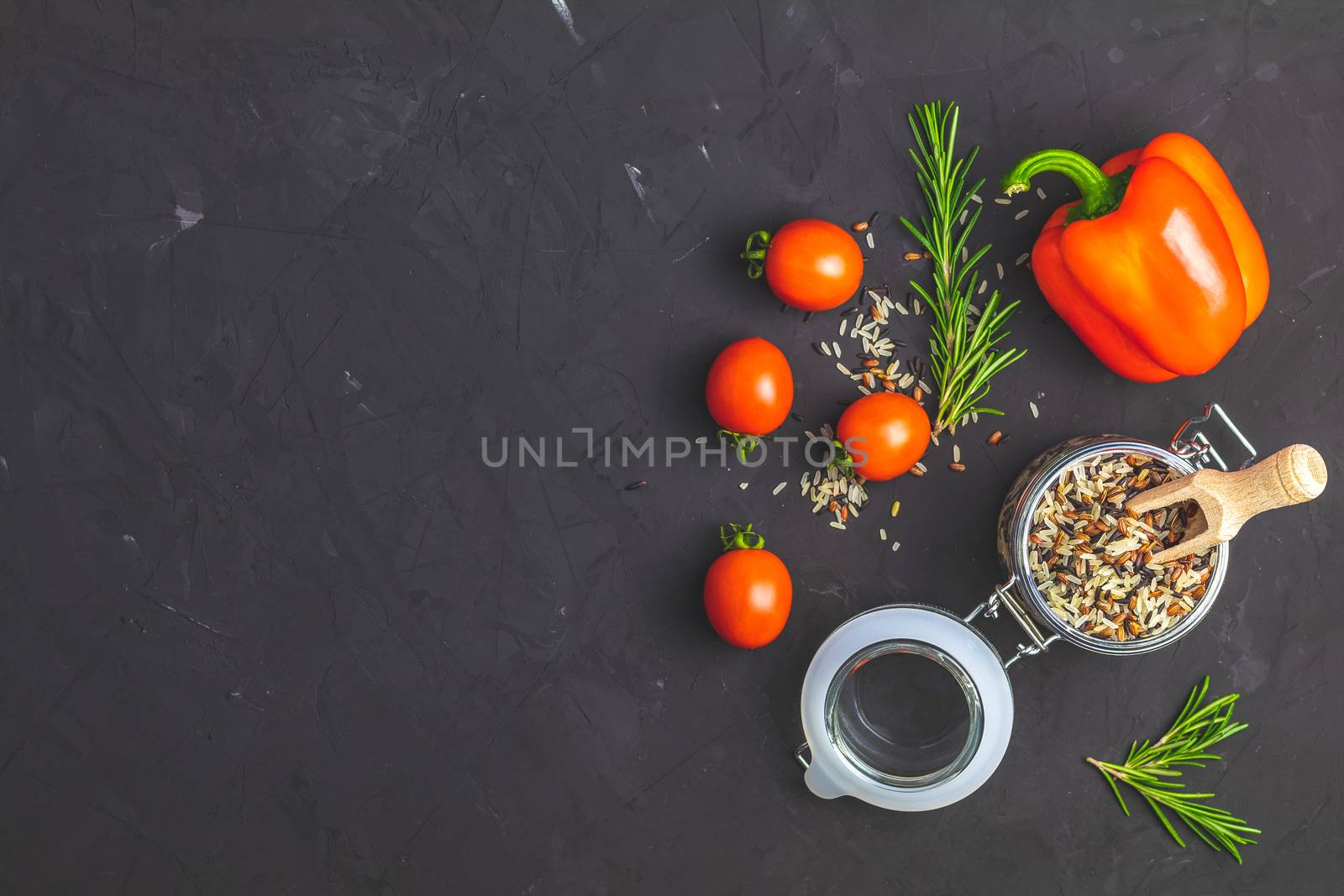 Black, purple, and white rice (Oryza sativa) mix in glass jar on black stone concrete textured surface background. Raw pepper, tomatoes, olive oil and rosemary bunch. Top view with copy space for your text.