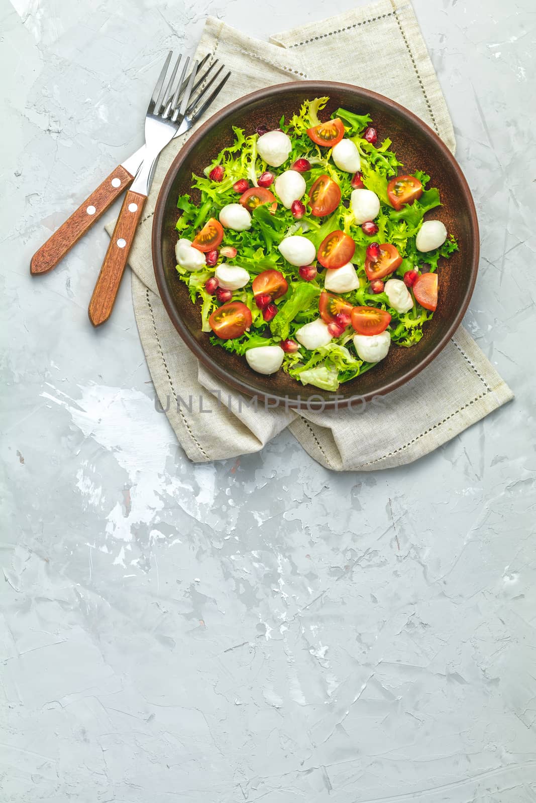 Fresh Cherry Tomato, Mozzarella salad with green lettuce mix served on a brown ceramic plate, healthy food, light gray stone concrete surface, top view, copy space.