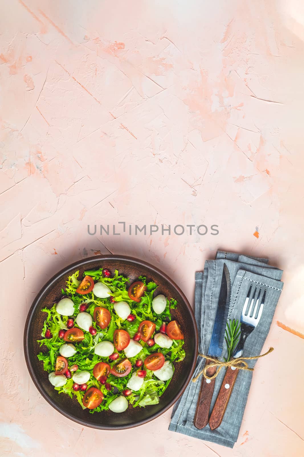 Fresh Cherry Tomato, Mozzarella salad with green lettuce mix served on a brown ceramic plate, healthy food, light pink living coral stone concrete surface, top view, copy space.